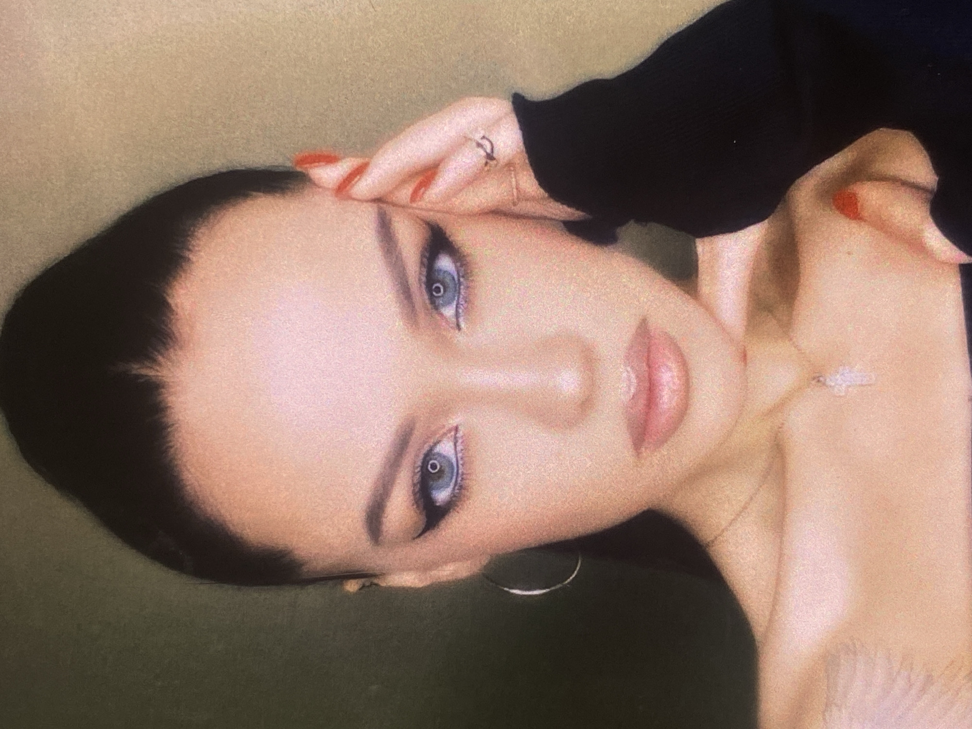 An extreme close-up portrait shot shows a young woman with dark hair pulled back into a high ponytail. She has striking blue eyes, accentuated by dramatic black winged eyeliner. Her lips are slightly parted, and she wears a delicate cross necklace. Her left hand rests on her temple, adorned with rings, while her right hand is visible with bright red nail polish. She is wearing a black off-the-shoulder top. The background is a blurred, muted green-yellow color. The image is grainy and has a soft focus effect. There is no discernible city or location information in the image.