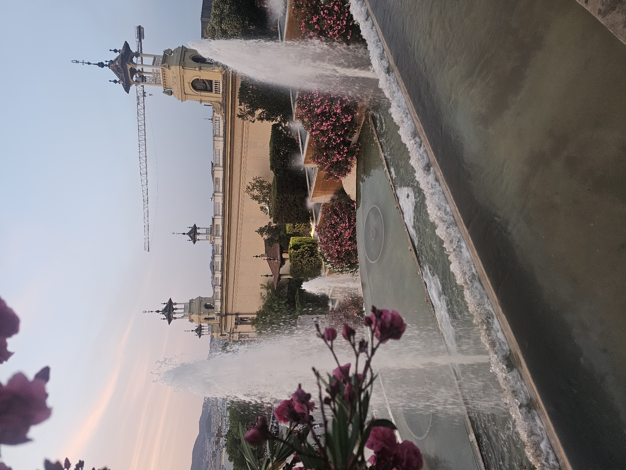 This image captures a scenic view of the Magic Fountain of Montjuïc in Barcelona, Spain. In the foreground, several tiered fountains are in operation, with water cascading down and creating a misty spray. Lush green bushes and vibrant pink flowers surround the fountain's edges, adding color to the scene. In the midground, a large, cream-colored building with ornate architectural details and towers stands prominently. A construction crane is visible in the background, suggesting ongoing development in the area. The sky above is a soft blend of pink and pale blue, indicating either sunrise or sunset. The overall atmosphere is serene and picturesque.