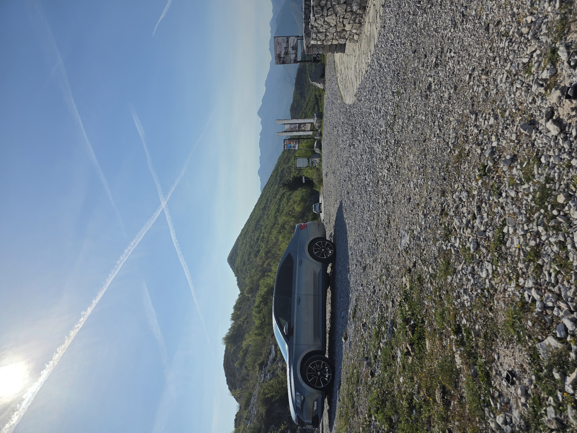 The image shows a gray Skoda Octavia car parked on a gravel surface. The surface is mixed with patches of grass and vegetation. In the background are mountains covered in green trees and distant mountain ranges under a clear blue sky with contrails. The location is a scenic overlook in Arbnež, Montenegro, marked by informational signage and a low stone wall at the edge of the overlook. The sun is visible in the lower-left corner of the image.