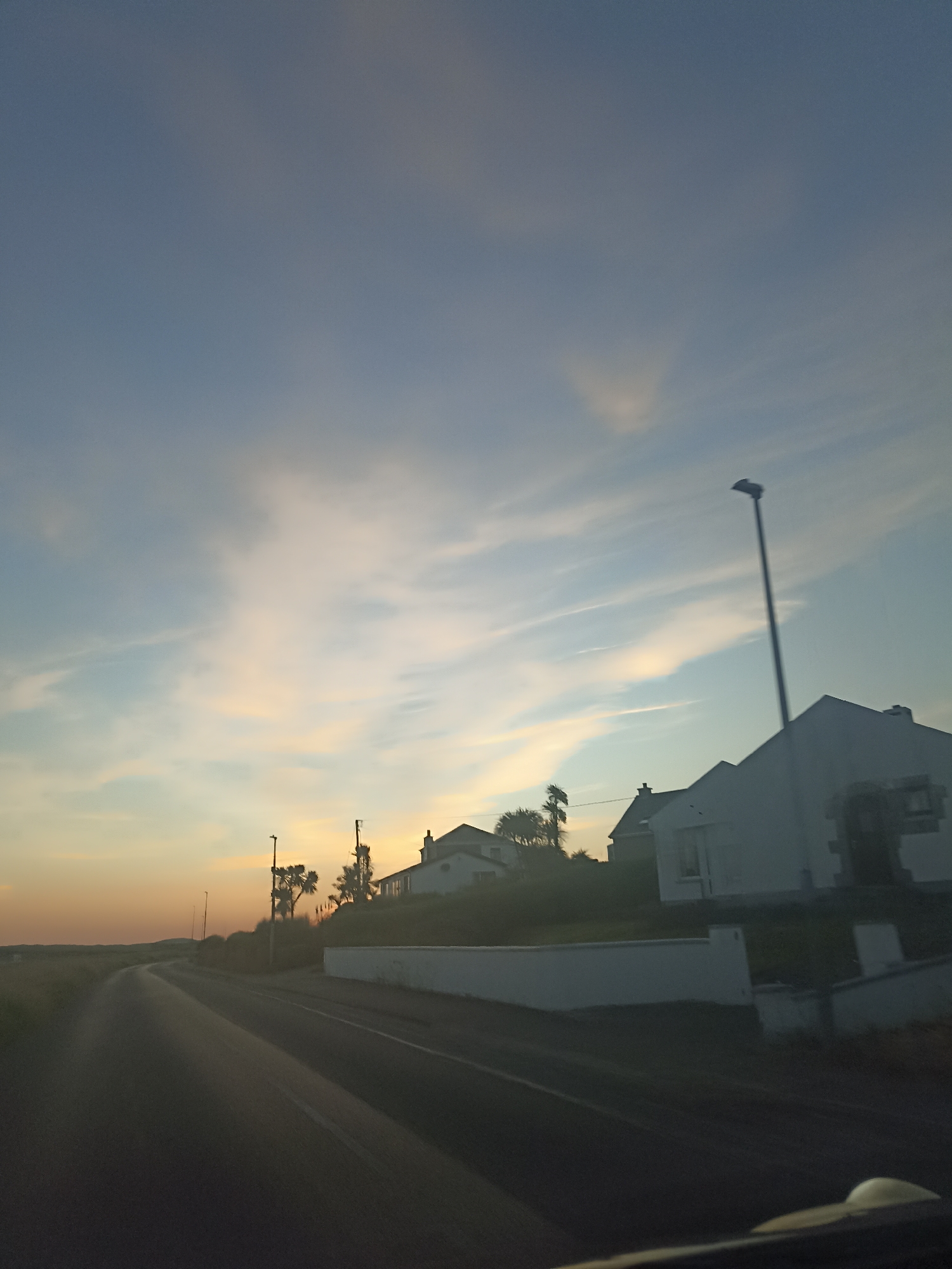 This is a photograph of a coastal road in Ireland at dusk. The sky is a blend of blue and orange, with wispy clouds illuminated by the setting sun. Several white houses are visible on a grassy hillside overlooking the road. A tall lamppost stands near one of the houses, and there are some trees and utility poles in the background. The road itself is paved and appears to curve into the distance. The overall impression is one of a tranquil, rural evening scene.