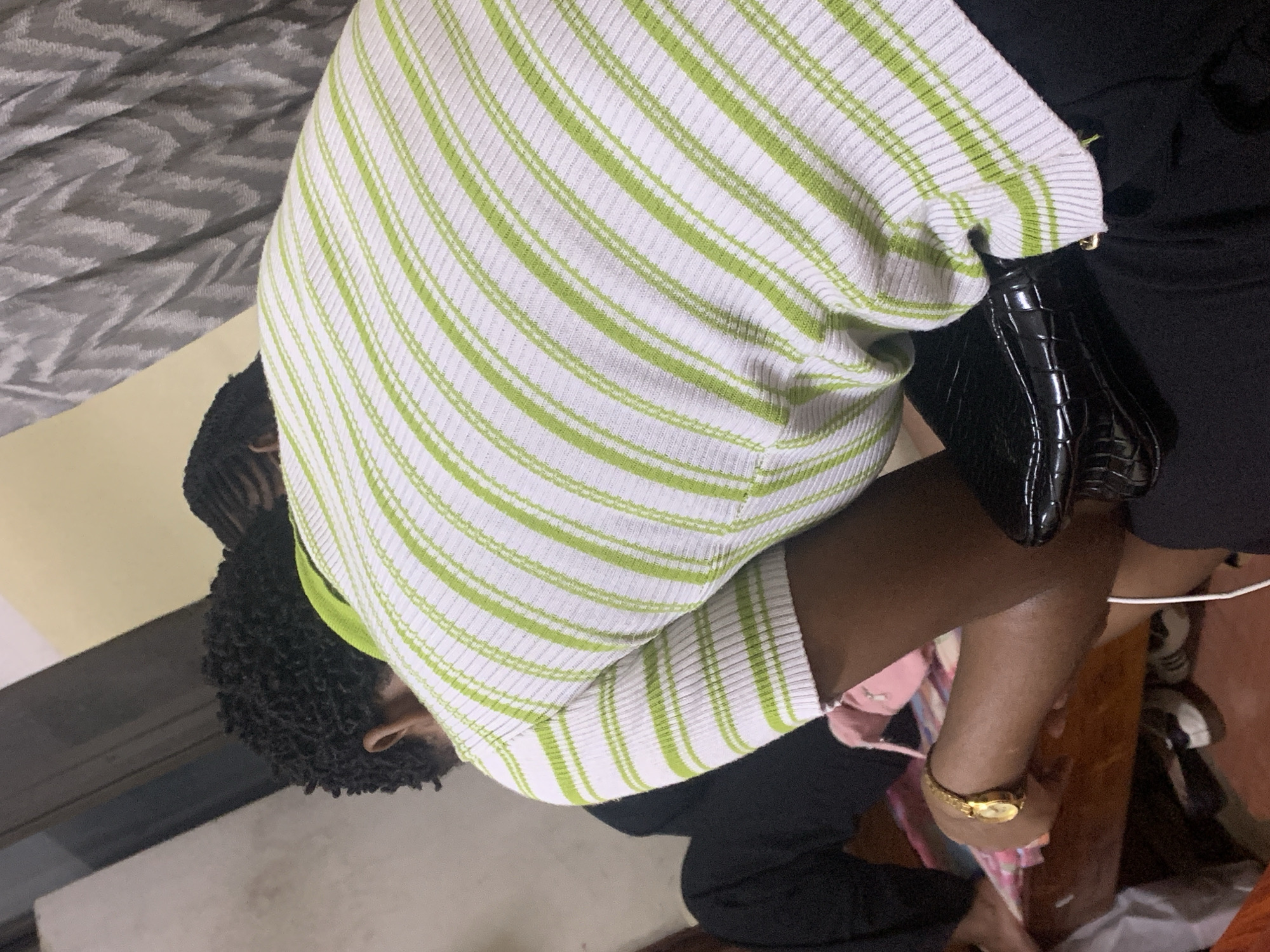A person wearing a white and green striped shirt is sitting down. They are holding a black purse. The person is also wearing a gold watch. The location cannot be determined from the image.