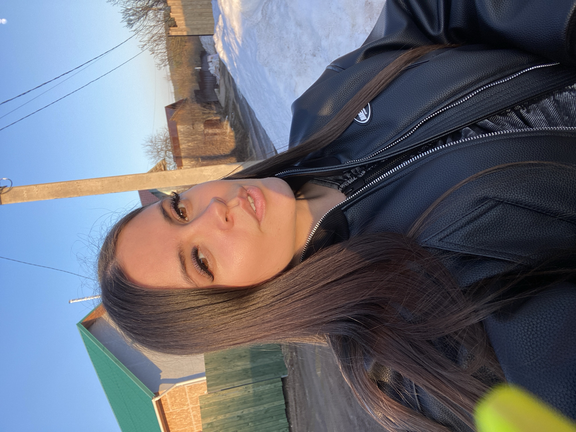 A medium shot captures a woman with long, straight brown hair and light skin. She is wearing a black leather jacket with a visible zipper. The background reveals a rural environment with a house featuring a green roof and tan siding. Snow is visible in the upper portion of the frame, suggesting a colder climate. A wooden fence is also present. The photograph was taken in Прикамский, Russia.