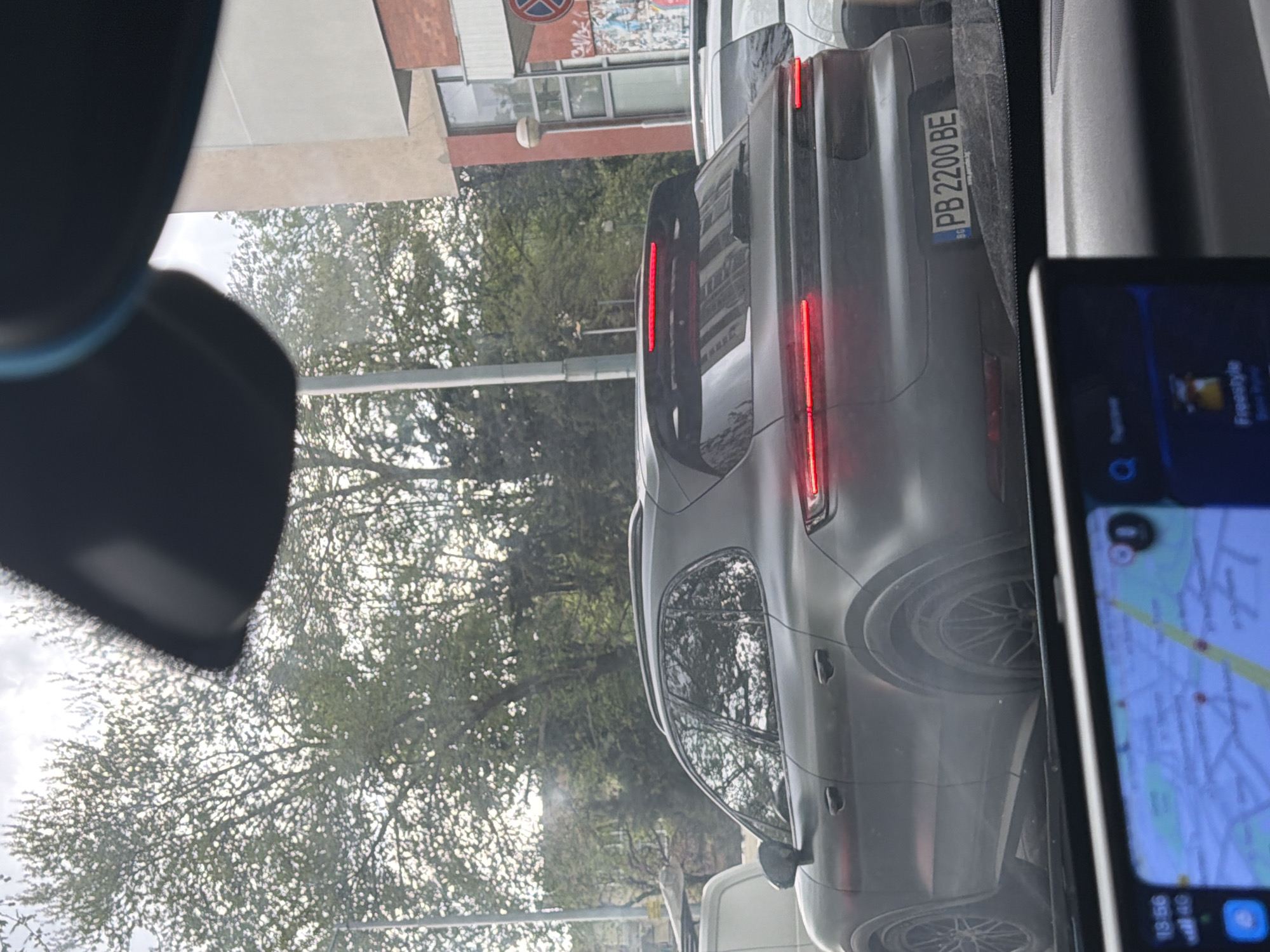 The image captures a street scene from the perspective of someone inside a vehicle, looking ahead at the rear of another car, likely stopped in traffic. The primary subject is a dark grey SUV, possibly a Citroën C5 Aircross, with its distinctive vertical red LED taillights illuminated.
A key notable detail is the SUV's license plate, which reads "PB 2200 BE". The "PB" designation, along with the "BG" country code visible below it, identifies the vehicle as registered in Plovdiv, Bulgaria.
To the right, a smartphone or tablet is partially visible, displaying a navigation application with a street map. The screen also shows a timestamp of "13:56" and a "4G" network indicator, suggesting the photo was taken in the early afternoon. Below the map, a music player interface is visible, showing the text "Freestyle".
The background features trees with bright green foliage, possibly indicating spring or summer, under what appears to be an overcast or bright sky. Hints of buildings are visible through and behind the trees. Parts of the viewer's car interior, such as a headrest or dashboard, are visible on the left side of the frame. The activity taking place is likely waiting in traffic, given the illuminated taillights.