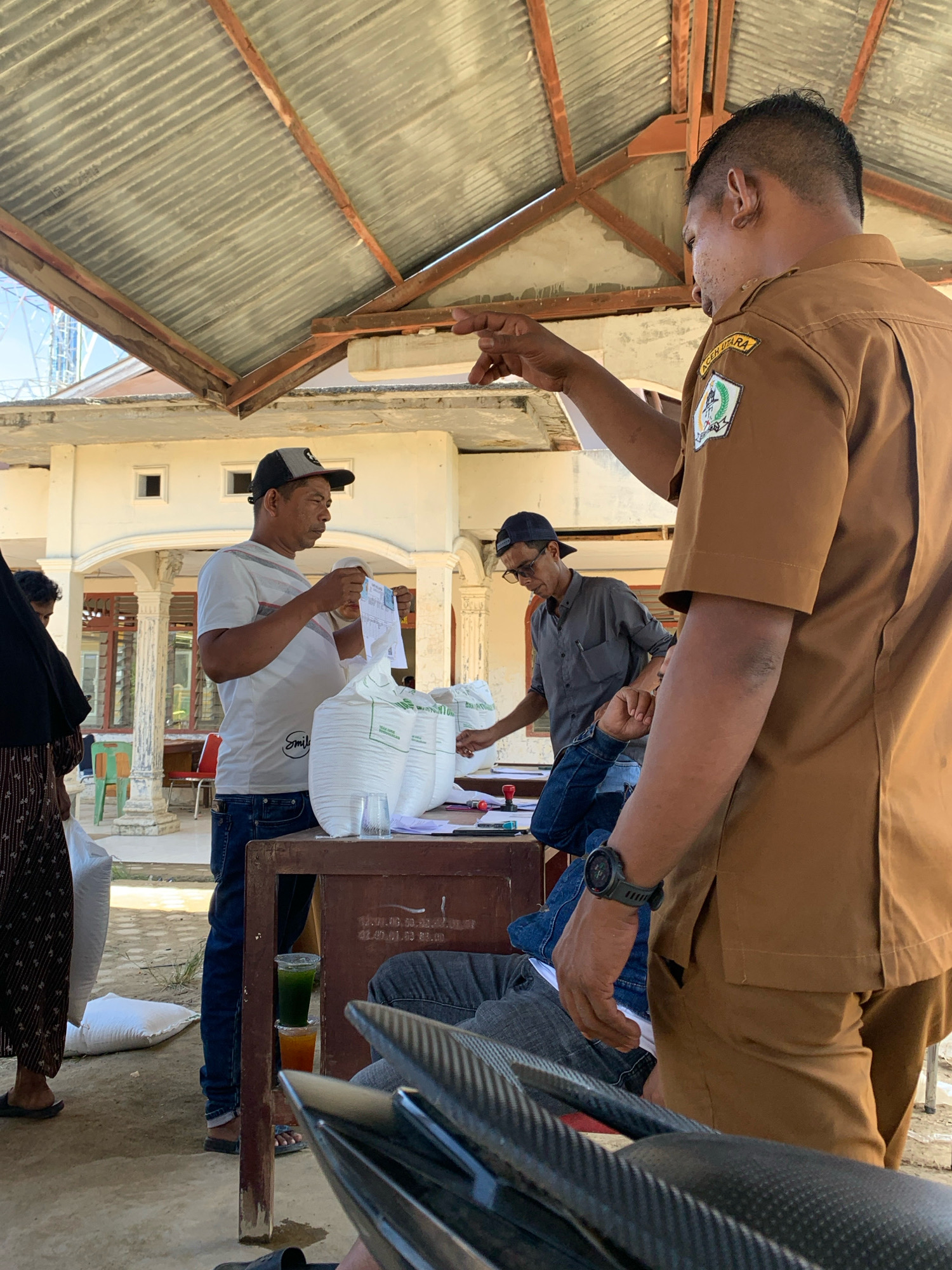 In Pantegurah, Indonesia, under a corrugated metal roof supported by wooden beams, a group of people are gathered around a table. A man in a brown uniform, possibly a government official, gestures with his right hand towards a table where another man in a white t-shirt and baseball cap is holding a piece of paper. Several large white sacks, likely containing goods, are stacked on the table and the ground. The man in the brown uniform has a patch on his left shoulder with an emblem and text that reads "ACEH UTARA". Another man in a gray shirt and a dark baseball cap is leaning over the table, looking at documents. The environment appears to be an outdoor or semi-outdoor setting, with a building visible in the background. The ground is dirt and gravel. The scene suggests an administrative or distribution process.