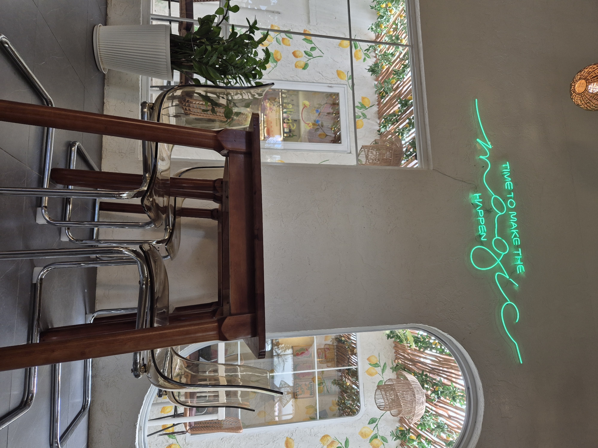 The image depicts an indoor cafe with a bright, airy ambiance, featuring a prominent neon sign on a textured wall that reads "TIME TO MAKE THE MAGIC HAPPEN" in green lettering. Beneath the sign, a wooden table is set with four clear chairs with chrome legs. To the left, a potted plant in a white ribbed pot sits on a dark tiled floor. The background reveals windows with arched openings, adorned with lemon motifs and bamboo-like structures, suggesting an outdoor or garden-themed aesthetic. Woven pendant lights hang from the ceiling, contributing to the decorative atmosphere. The scene appears to be a well-lit, aesthetically designed space, possibly in a cafe or restaurant located in Da Nang, Vietnam, given the information provided.