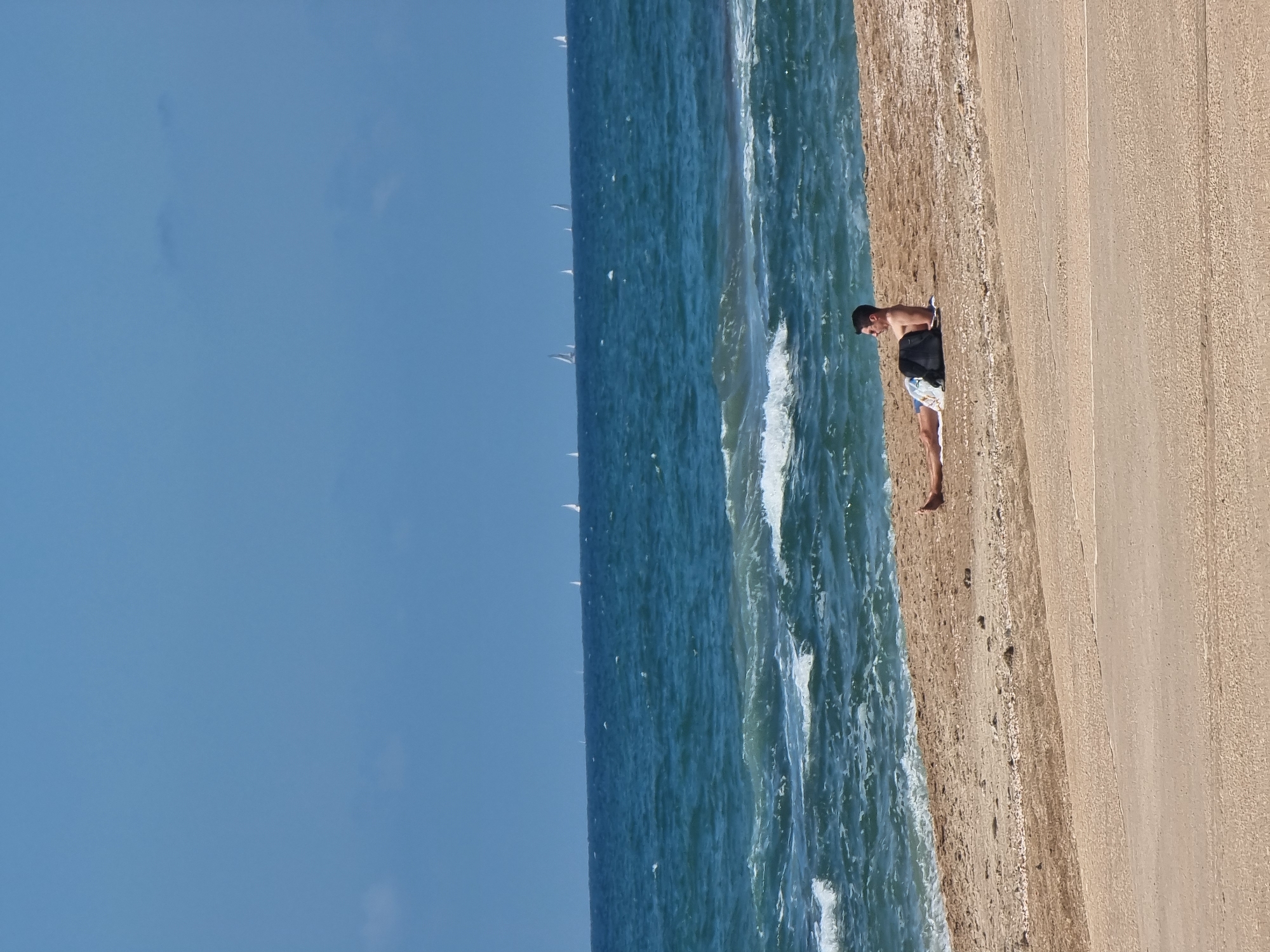A person is standing at the edge of the beach, in the city of Tel-Aviv, Israel. The image captures the person wearing swimming trunks. The scene includes a sandy beach, the sea with waves, and the sky in the background.