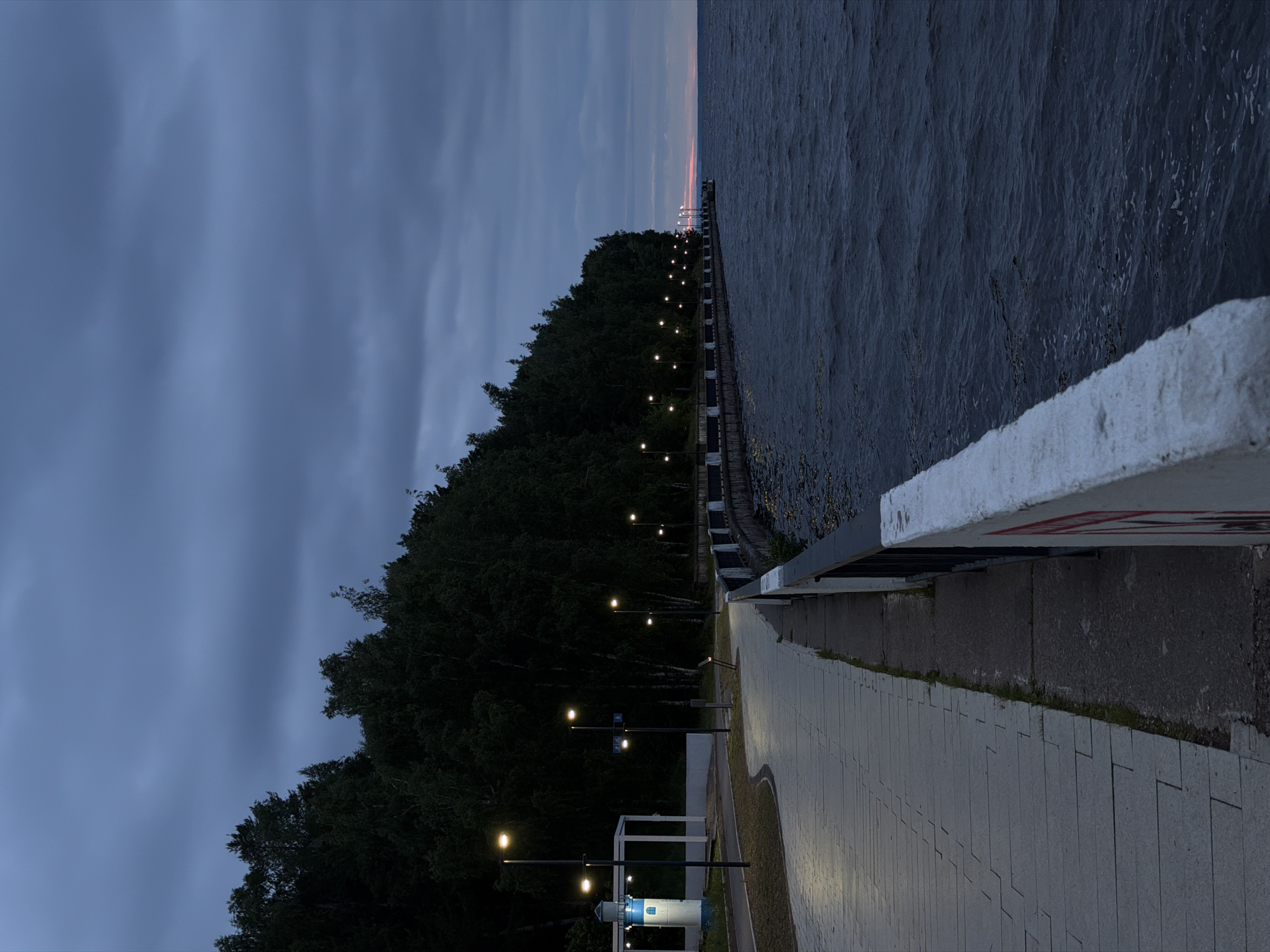 This image depicts the Kama River embankment in Chaikovsky, Perm Krai, Russia, during twilight or early night. The sky is heavily overcast with dark blue-grey clouds. A paved promenade, constructed from light-grey rectangular pavers, begins in the mid-left foreground, curving to the left before straightening into the distance. Along the right side of the promenade, a white concrete barrier with a dark metal railing separates the walkway from a wide body of dark, choppy water, consistent with a large river or reservoir. The barrier shows signs of wear and a partially visible red marking in the lower right. A series of tall, dark streetlights with warm yellow illumination are evenly spaced along the promenade, casting light onto the path. To the left of the walkway, a dense line of dark-foliaged trees, likely birches and other deciduous species, forms a forest edge running parallel to the embankment. In the mid-left, a small, decorative structure resembling a lighthouse, white with a blue top and small windows, stands near the promenade. In the far distance, a subtle reddish-orange glow from the setting sun is visible on the horizon, just above the water. The water exhibits small waves and ripples. There are no individuals present in the scene.