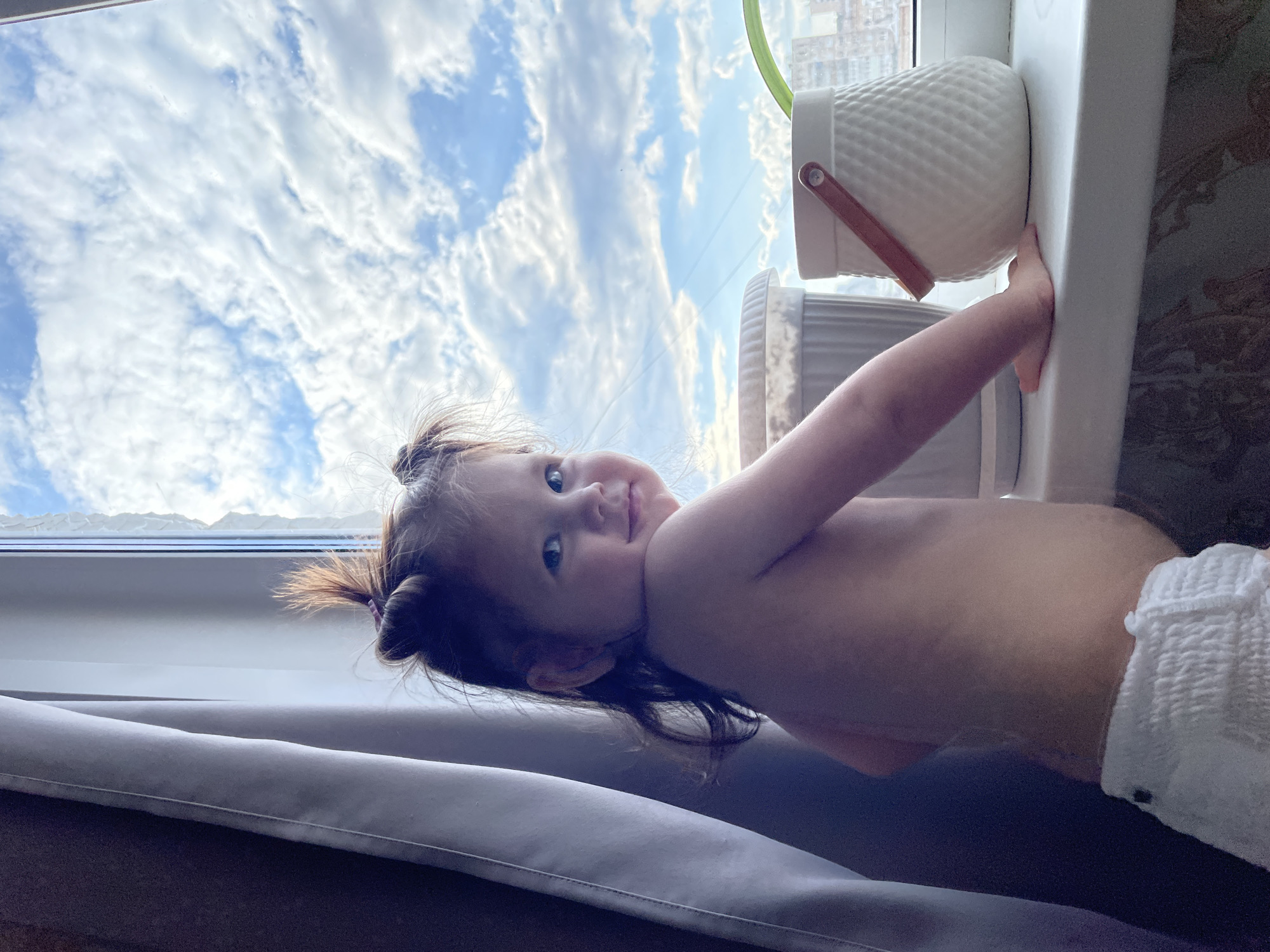 A young child, likely a toddler, is depicted in an indoor setting, leaning against a white windowsill. The image is rotated 90 degrees counter-clockwise from its upright orientation. The child has dark hair styled in two small buns or pigtails and is shirtless, wearing a white diaper. Their left arm is extended towards a white, textured planter with a brown handle or strap, which rests on the windowsill. The child looks directly at the camera with a calm, curious, and slightly smiling expression.
Through the large window behind the child, a bright blue sky filled with numerous white, fluffy clouds is visible, indicating a sunny daytime. Faint outlines of distant buildings suggest an urban environment, consistent with Izhevsk, Russia. Below the child, a light grey curtain or cushion is visible, providing a soft surface. There is no visible text in the image.