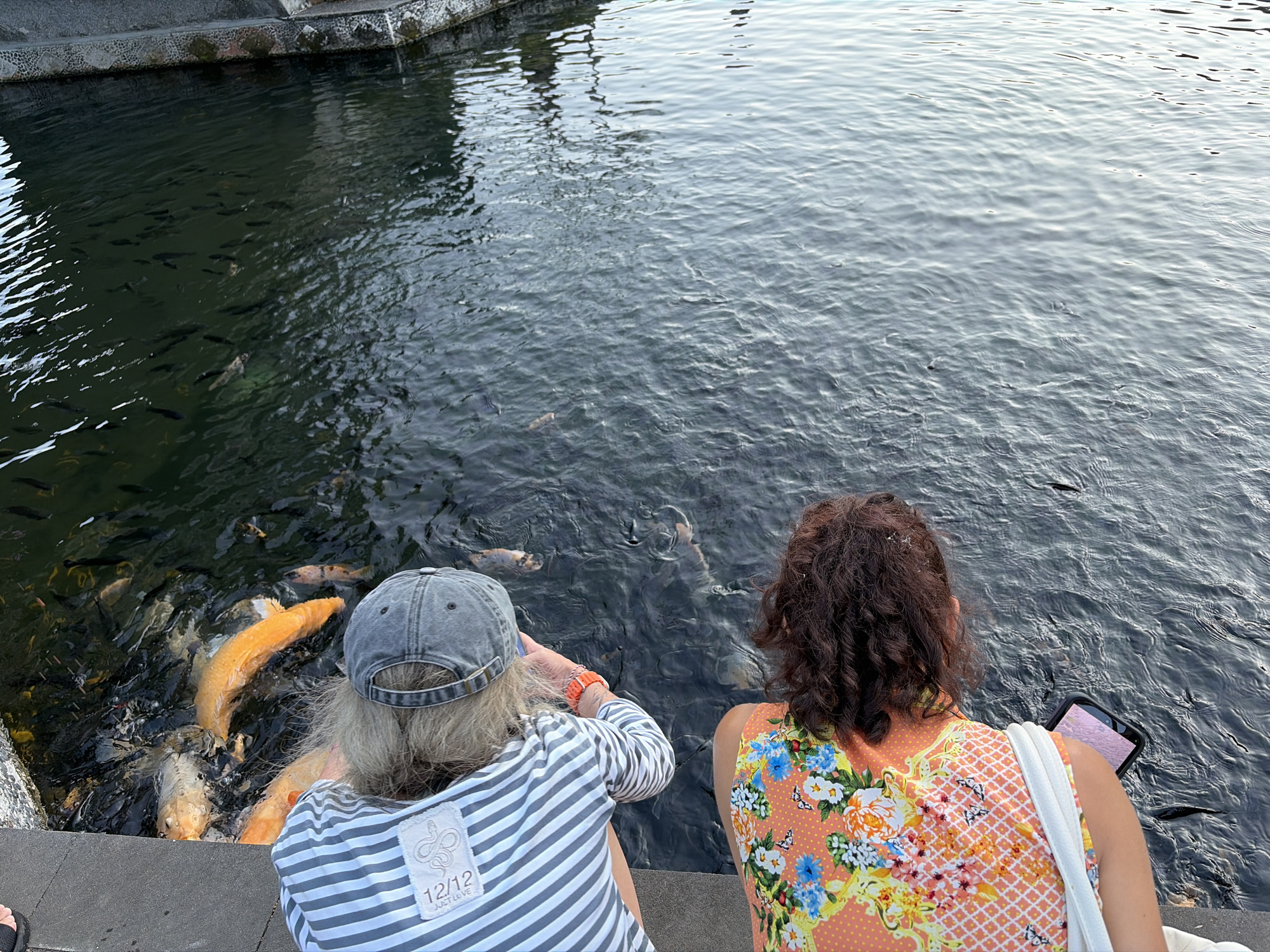 Two women are sitting by a body of water, likely a pond or canal, in Abang, Indonesia, with many fish swimming near the edge. The woman on the left, wearing a grey baseball cap and a striped shirt, is leaning towards the water, her hand extended. She has long, greyish-blonde hair. The woman on the right, with curly brown hair, is wearing a vibrant orange top with a floral pattern. She is holding a tablet in her lap. The water is dark and slightly rippled, with numerous fish visible, including several large, orange koi. The edge of the water is lined with a grey, concrete structure. The lighting suggests it might be late afternoon or early evening.