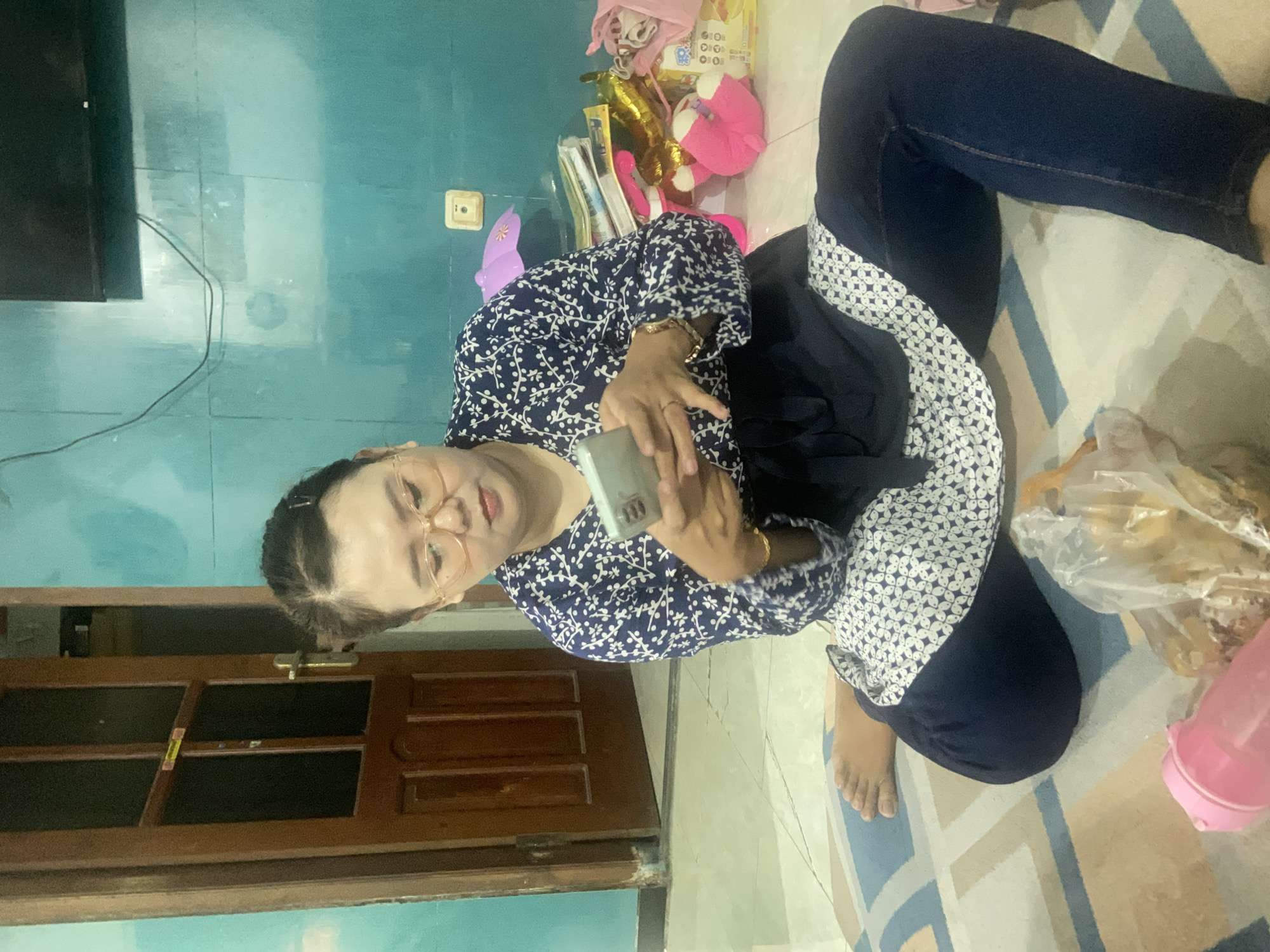 A woman is seated on the floor indoors, holding and looking at a smartphone. She has light skin, wears round-rimmed glasses, and her hair is pulled back. She is dressed in a dark blue long-sleeved shirt with a white floral pattern, blue jeans, and another dark garment with a white geometric pattern around her lap. A ring is visible on her right hand, and a gold-colored bracelet on her right wrist.
The setting appears to be a casual living space within a home in Gelam, Indonesia. The walls are a distinct teal or light blue color. The floor is covered by a patterned rug featuring geometric shapes in shades of blue, beige, and white. To the left, a dark flat-screen television is mounted on the wall, with a black cable hanging down. A light-colored electrical outlet is visible on the teal wall. In the lower-left background, a wooden door, partially open, shows a glimpse of another area.
To the right of the woman, a clear plastic bag containing what appears to be snacks or food items, and a pink plastic bottle are resting on the rug. The background behind the woman reveals shelves or a surface with various items, including pink stuffed animals, gift bags, and other household clutter. The overall scene suggests a relaxed, everyday moment indoors, likely during the daytime given the ambient lighting. There is no discernible text visible on any objects in the image.