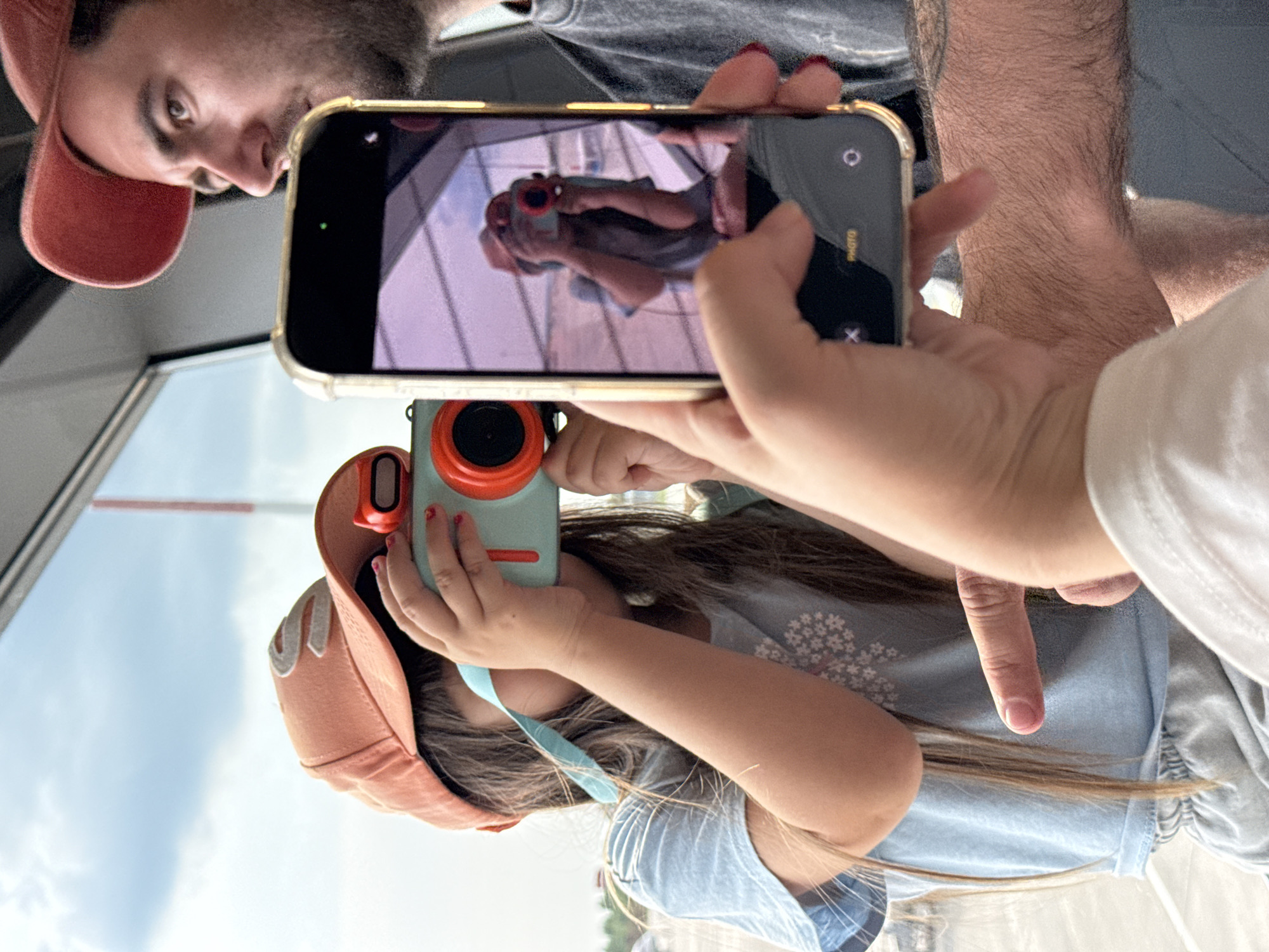 In a sunlit interior, likely an airport terminal given the large window and sky visible, a young girl wearing an orange cap and a light blue t-shirt is focused on taking a photograph with a teal and orange toy camera. Her hands are positioned to operate the camera, and a turquoise strap hangs from it. To her right, a man wearing a red cap and a dark t-shirt is looking towards the girl, with his arm held out holding a gold-rimmed smartphone. The phone's screen displays a reflection of the girl taking a picture with her toy camera, creating a layered photographic moment. The environment outside the window appears to be an airfield or open area with a cloudy sky. The perspective suggests the photo is taken from a third person's viewpoint, possibly a parent, capturing this interaction. No specific landmarks identify the location as Bol'shie Kabany, Russia.
