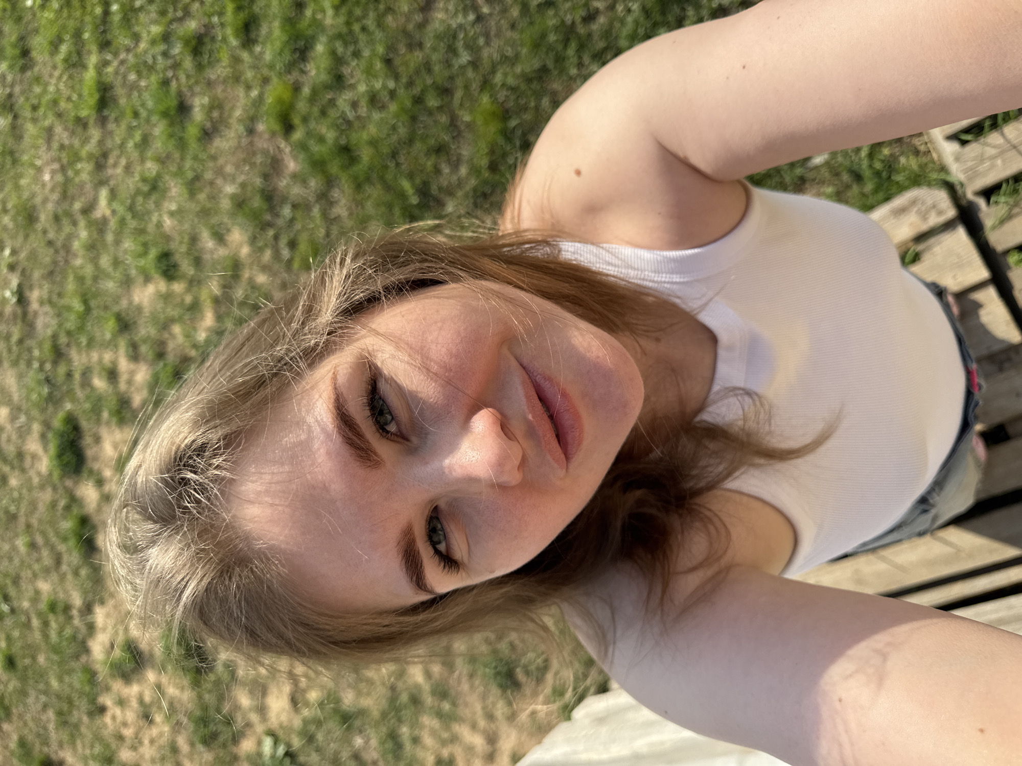 A young woman in a white tank top is posing for a selfie outdoors. The background shows grass and wooden planks. The location is Ферма, Russia.