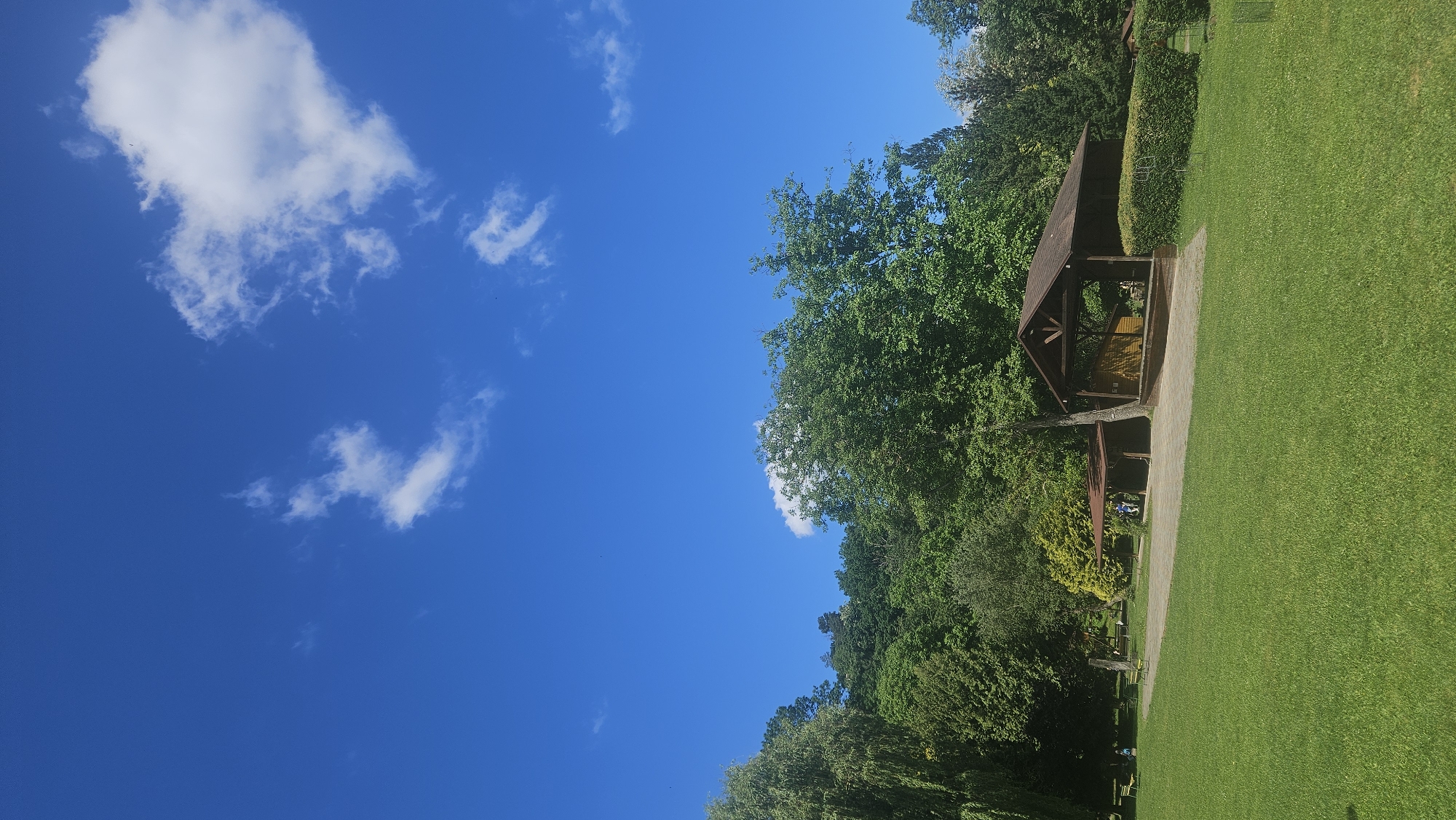 The image depicts a sunny day in a park-like setting with a clear blue sky and scattered white clouds. In the midground, a wooden gazebo structure is visible, surrounded by lush green trees and grass. There is likely an area of lawn, manicured grass, or parkland. The location is in Prešov, Slovakia, based on the coordinates provided.