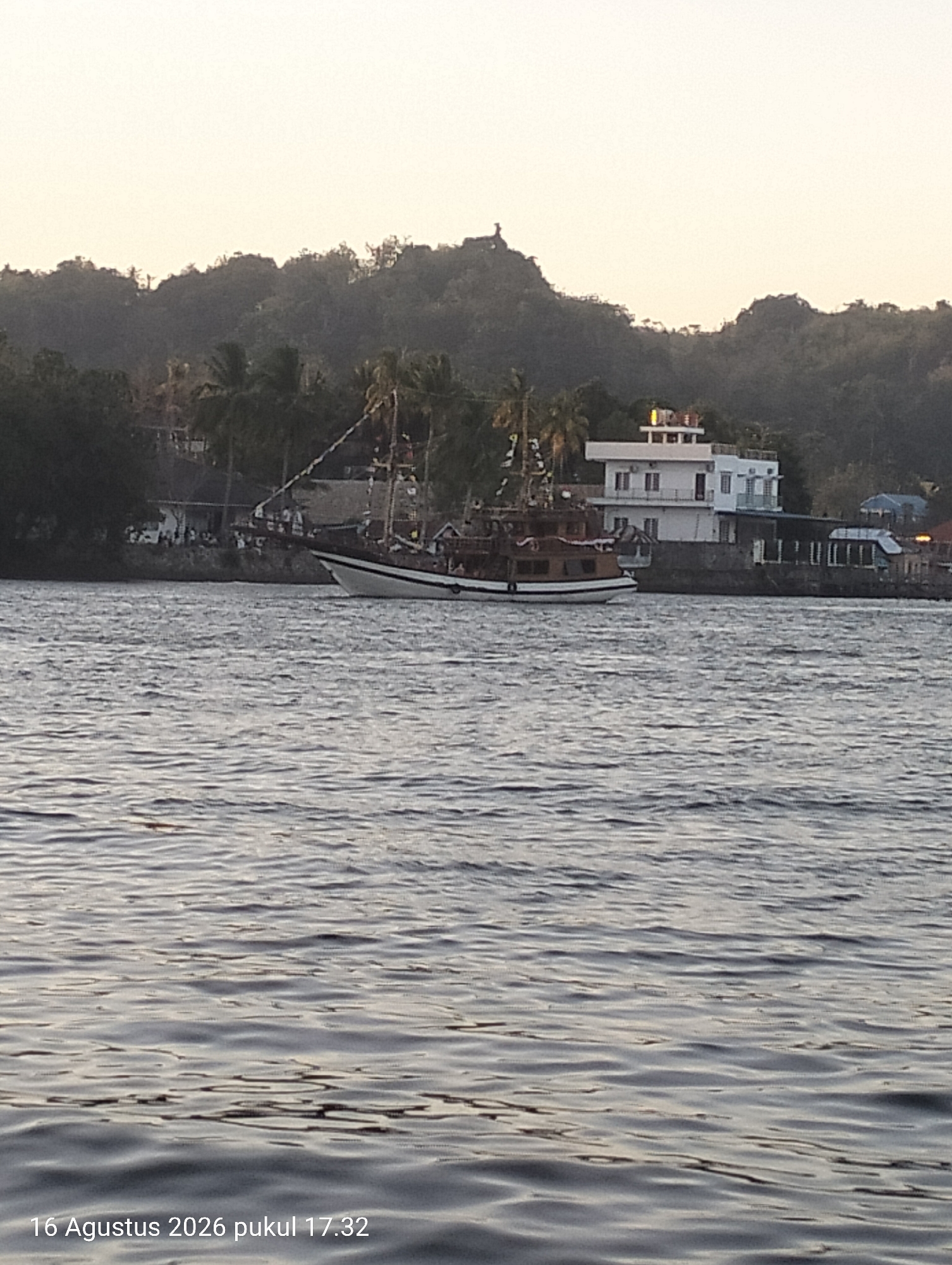 This image, captured on August 16, 2026, at 17:32, shows a scene likely in Kendari, Indonesia, featuring a body of water, a boat, and a shoreline with buildings and lush vegetation.
The foreground is dominated by the water, which has a rippled surface, reflecting the subdued light of what appears to be late afternoon. Several dark, horizontal lines of shadow and lighter, wavy patterns suggest moderate chop.
In the middle ground, a large wooden boat with white trim is positioned near the shore, appearing to be at anchor or moving slowly. The boat has multiple masts and rigging, suggesting it might be a schooner or a similar sailing vessel. The hull is a dark brown or red-brown color.
The shoreline is lined with trees, including palm trees, and several buildings. The most prominent building is a white, multi-story structure with balconies and a flat roof. There are other, smaller buildings visible further along the shore, some with darker roofs. A group of people can be seen gathered on the shore near the boat, though their details are indistinct.
The background consists of a dense, forested hillside that rises steeply behind the shoreline buildings. The vegetation is dark green, and the contours of the hill are softened by distance and possibly haze. The sky above is a pale, washed-out yellow or cream color, indicating either a hazy atmosphere or the sun setting behind a cloud bank.
The overall impression is a coastal scene, likely a port or a bay, with a blend of natural landscape and human habitation. The time of day suggests a tranquil, possibly popular, location for leisure or transit.