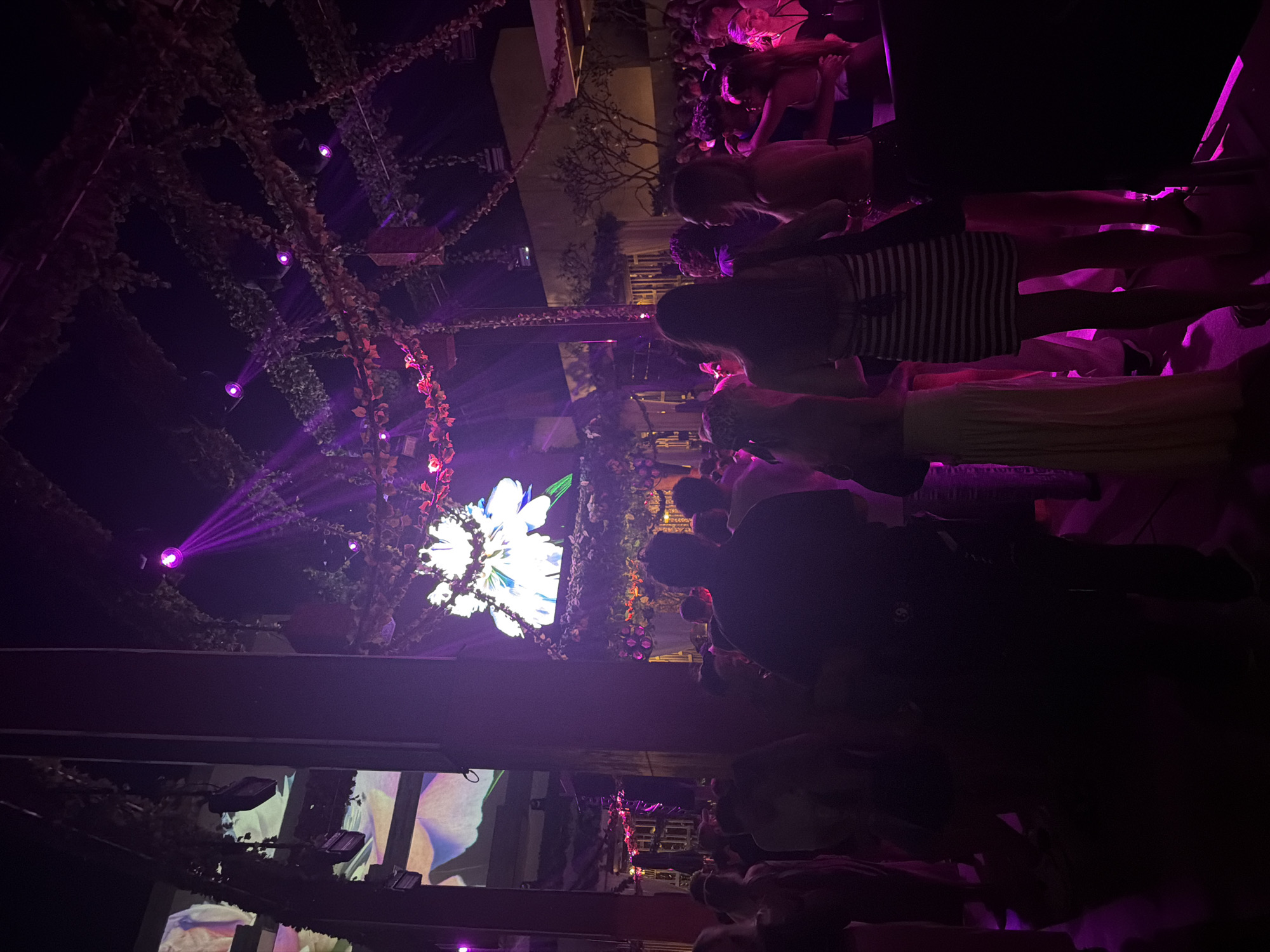 The scene is set at night in Kuta Selatan, Indonesia, at an outdoor venue decorated with floral arrangements and vine-covered structures lit with purple spotlights. A large flower-shaped light fixture is visible against the night sky. A crowd of people are standing and interacting in the space, suggesting a social event. Some of the attendees are wearing dresses. A video projection screen displaying large floral imagery is visible below the elevated platform.