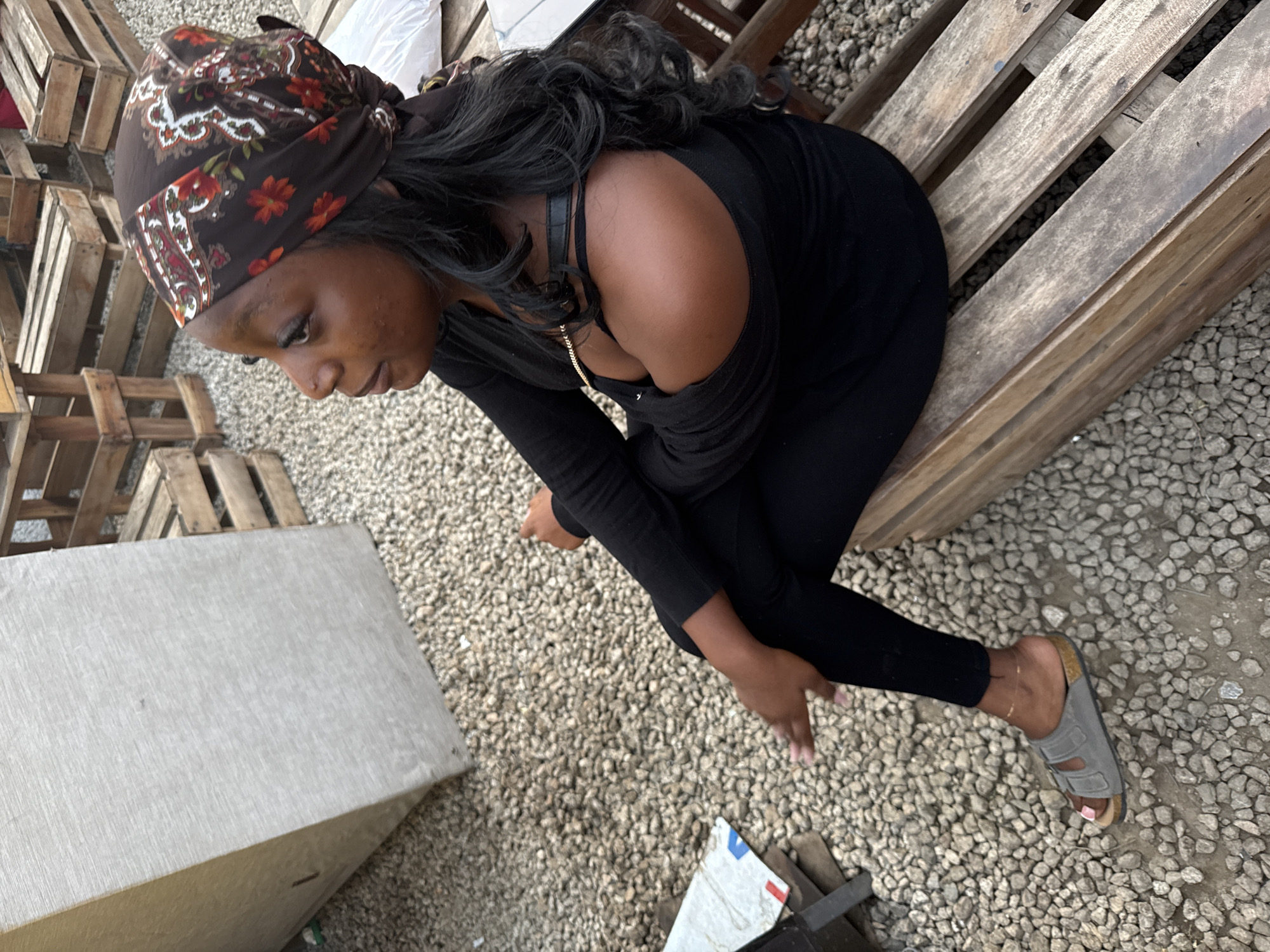 An overhead shot shows a Black woman with dark hair wearing a brown and red patterned headscarf and a black outfit with an off-the-shoulder design. She is sitting on a wooden pallet placed on a gravel surface. Her legs are bent and arms crossed, with her gaze directed downwards. She wears a gray sandal on her right foot, with her toes visible. In the background are stacked wooden pallets and a gray concrete block. The setting resembles an outdoor storage area or a construction site in Gwarinpa, Nigeria.