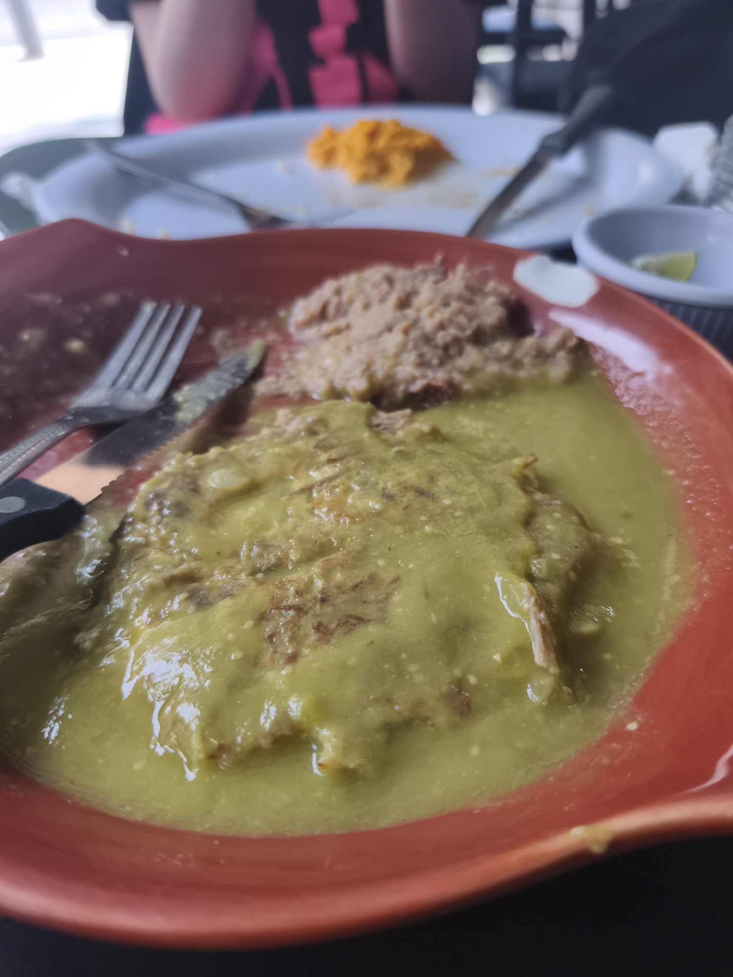 The image shows a partially eaten meal on a red plate. Refried beans and a dish covered in green sauce are visible on the plate. A fork and knife rest next to the food. In the background, another plate with remnants of orange food is visible. A small dish with a lime wedge is also present. The scene appears to be a casual dining setting, possibly a restaurant. The location is unknown, but the cuisine suggests it might be in Mexico City, Mexico.