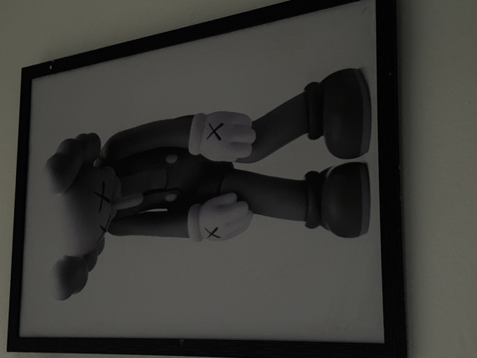 A framed black and white image of a cartoonish figure with large, round ears and X-shaped eyes is displayed on a wall. The figure wears overalls and large, round-toed shoes. The image is tilted, with the top left corner angled away from the viewer. The background appears to be a plain wall. The location is Ihiagwa, Nigeria.