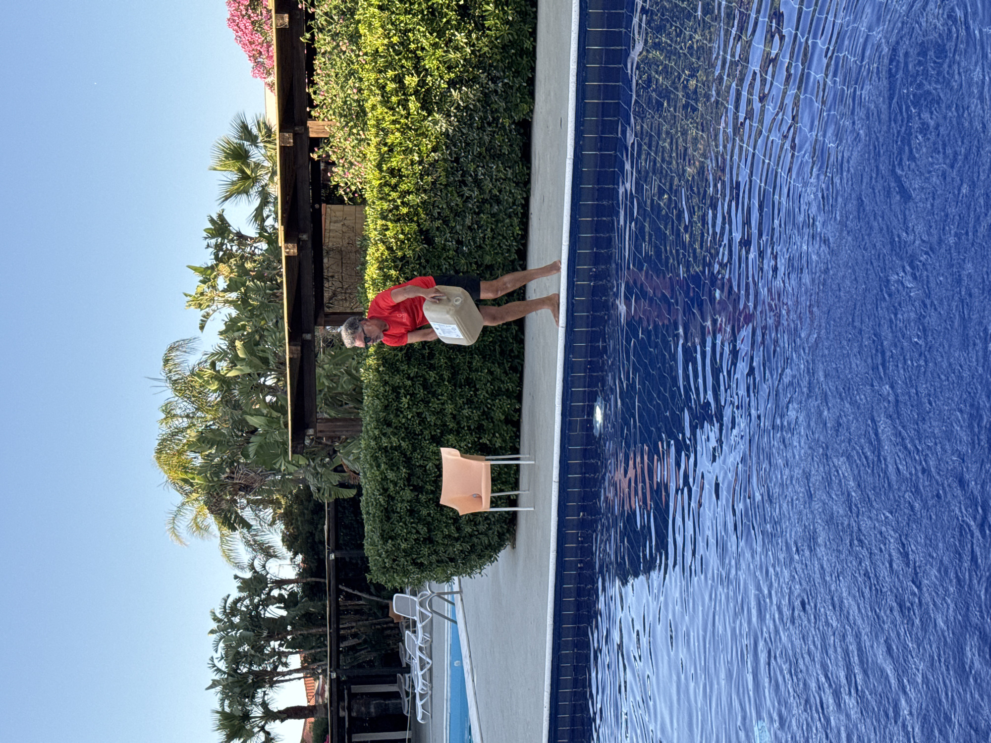 A middle-aged man with graying hair and sunglasses is standing barefoot on a concrete pool deck. He is wearing a red t-shirt and black shorts and is holding a large, light-colored plastic container with both hands. He is facing towards a swimming pool with blue tiles and is in the process of tilting the container as if to pour something into the water. To his left, there is an orange plastic chair. Behind him, there is a neatly trimmed green hedge, a wooden pergola, and palm trees under a clear blue sky. The location appears to be a resort or a private residence in Isola delle Femmine, Italy, given the presence of a swimming pool, lush vegetation, and architectural elements typical of the region. The lighting suggests it is either early morning or late afternoon.