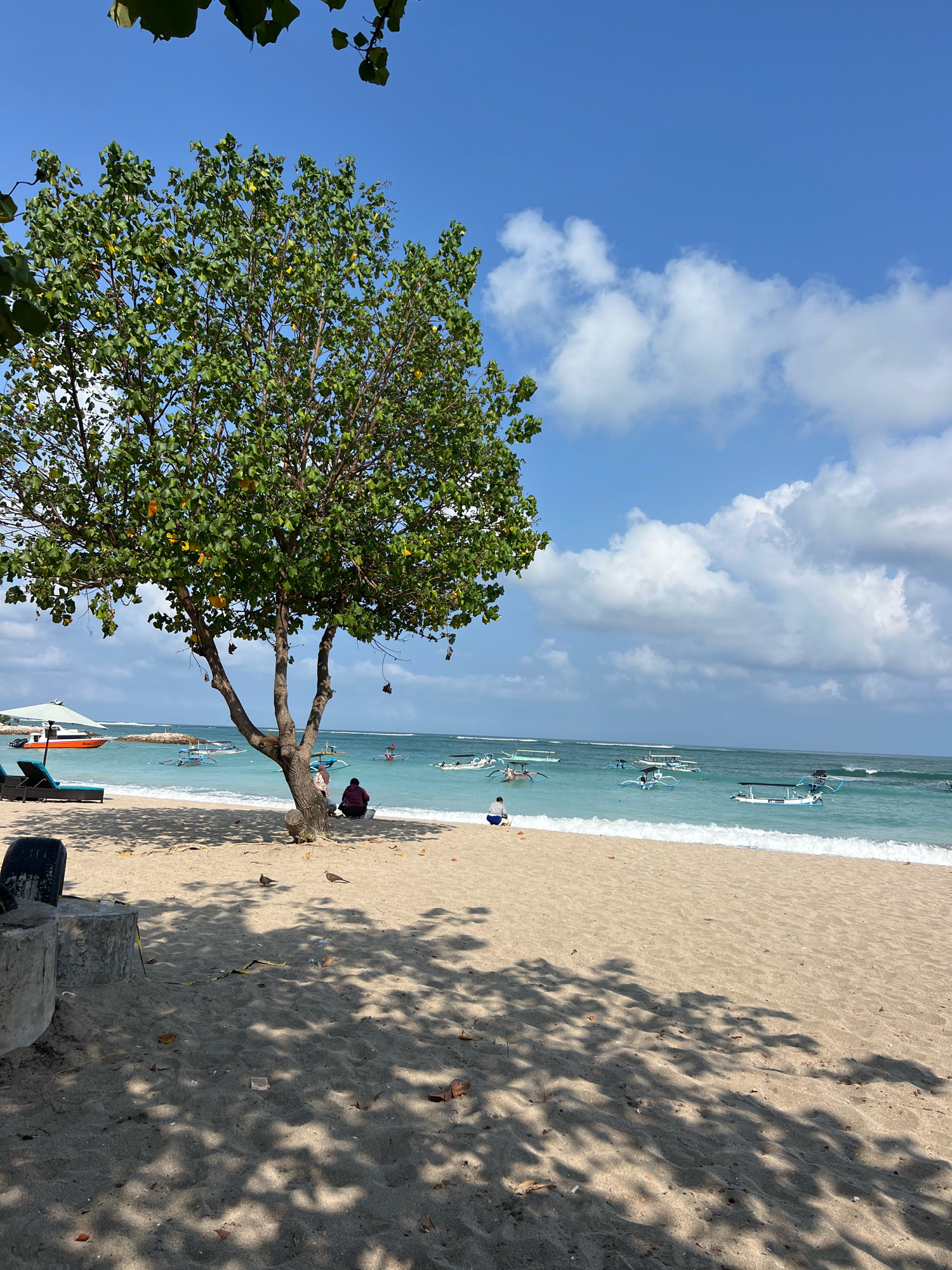 A full shot captures a tropical beach scene in Kuta, Indonesia. The foreground is dominated by sandy beach, with shadows from a tree cast across it. To the left, two concrete cylindrical structures and a discarded tire are visible. A lone tree with green foliage stands in the middle ground. Two people are seated beneath the tree, one in a maroon shirt and the other in white. Beyond them, the turquoise ocean stretches to the horizon, dotted with numerous small, traditional Indonesian fishing boats. A white umbrella and two lounge chairs are set up on the beach to the left. The sky is a clear blue, with scattered white clouds.