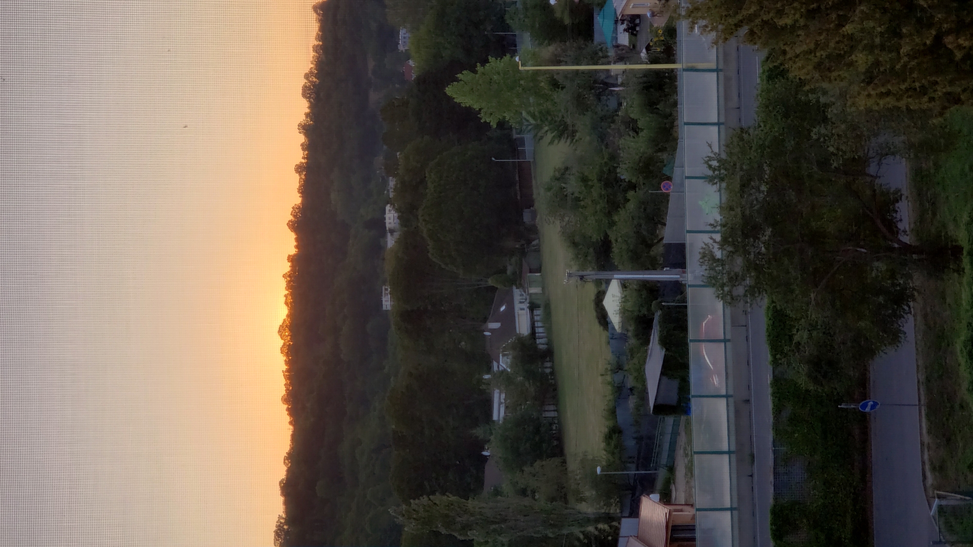 The image depicts a sunset over a tree-lined hill in the city of Prešov, Slovakia. The sky is a gradient of orange and pale yellow, with the sun just peeking over the horizon. Below the hill, a suburban landscape unfolds, featuring houses, buildings, and dense trees. In the foreground, a road is visible, bordered by a sound barrier and vegetation. A solitary tree stands in the middle ground. The scene is captured through a window screen, indicated by the fine grid pattern overlaying the image. There are no visible people or animals. The overall impression is of a peaceful, early evening urban environment.