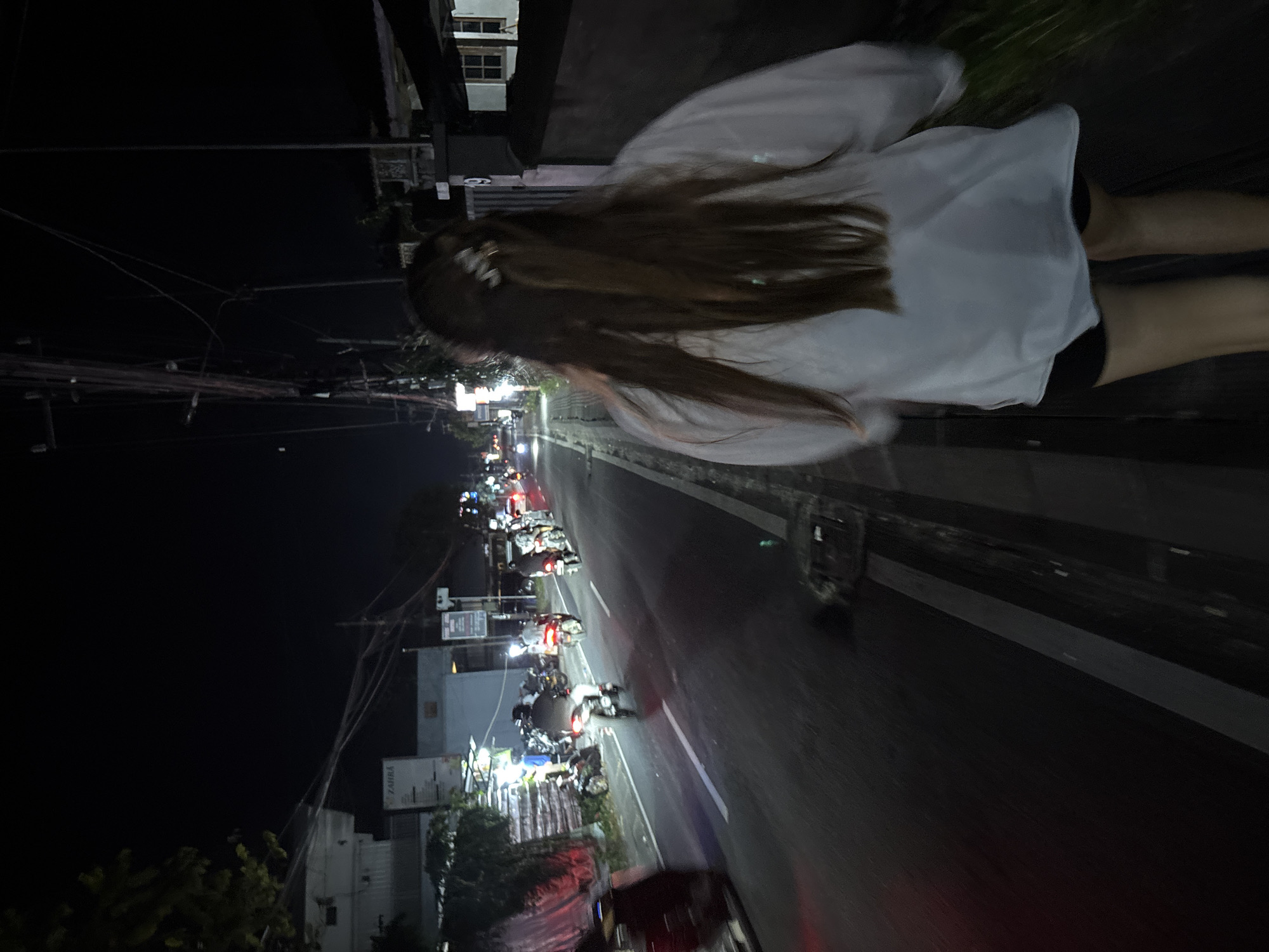 At night in Ungasan, Indonesia, a blurred image shows a person with long brown hair wearing a white top and black shorts, standing or walking on a sidewalk next to a road. The road is illuminated by streetlights and the headlights and taillights of numerous motorcycles and other vehicles. Buildings and electrical wires are visible on the left side of the road, receding into the distance. The scene is dark, with artificial lighting dominating the view.