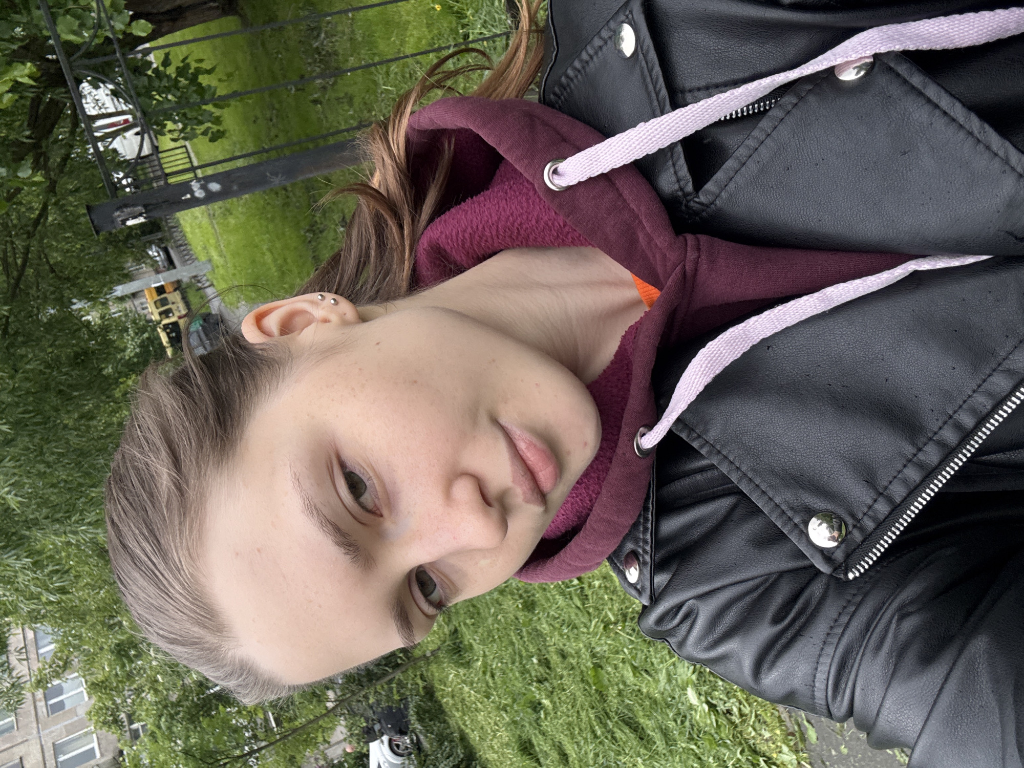 A young person with fair skin is pictured outdoors in Saint Petersburg, Russia. They are wearing a black leather jacket over a burgundy hooded sweatshirt with light pink drawstrings. The person has light brown hair and is facing slightly left of the camera. Visible in the background are green grass and trees, along with a black metal fence. A multi-story building is partially visible in the background, as well as some parked vehicles.