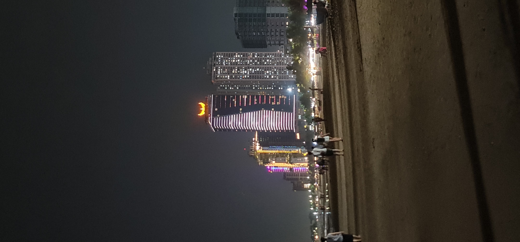 At night, the beach in Đà Nẵng, Vietnam is partially lit by the lights of the city. Several high-rise buildings, including one with a glowing "3" on top, line the coast. People stroll along the beach. A portion of the beach and the sky are dark.