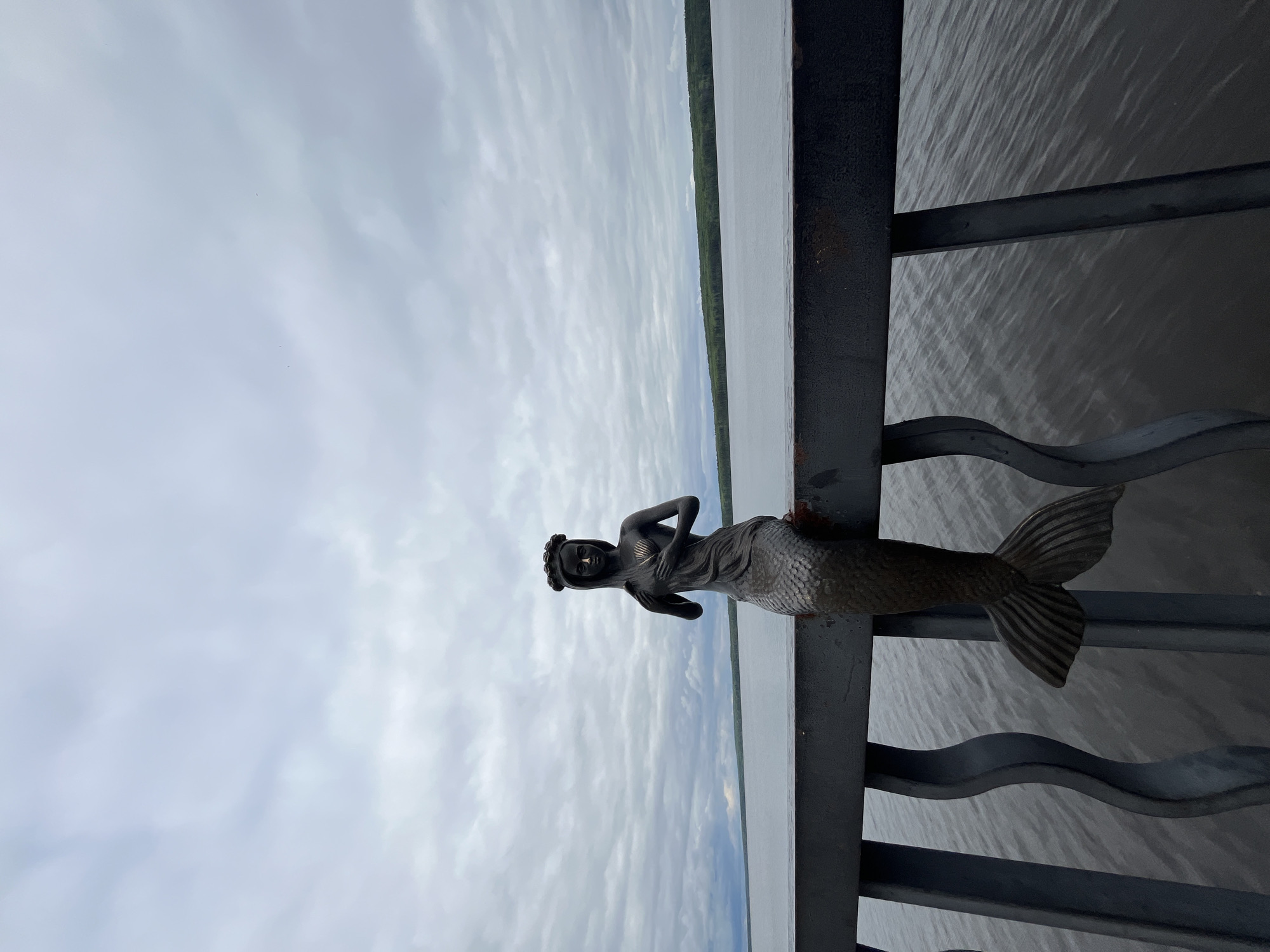 A statue of a mermaid is mounted on a dark grey metal railing. Beyond the railing is a body of water, with a thin strip of trees visible on the far shore. The sky is overcast with grey clouds. The image appears to be taken in a public location, potentially near a waterfront. The mermaid statue is detailed and seems to be made of bronze or a similar metal. Given the prompt mentioning the city of Chaikovsky, Russia, it can be inferred that this scene is likely located there.