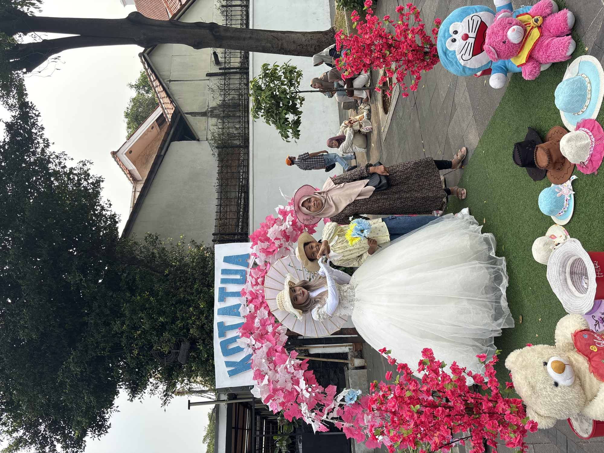 The image shows an outdoor scene in Kota Tua, West Jakarta, Indonesia. In the foreground, three individuals are posing in front of a decorative archway adorned with pink and white foliage and pink flowers. The person on the left is dressed in a white wedding gown and a straw hat, holding a white umbrella. To their right is a child wearing a yellow patterned shirt and a straw hat, holding a bouquet of blue and yellow flowers. The third person, standing to the right of the child, wears a light brown hijab and a dark patterned dress, holding a black purse.
Scattered on a green carpeted area in front of the archway are several hats, a large white teddy bear holding a red heart, and a blue Doraemon plush toy with a pink teddy bear. In the background, a white banner with the word "KOTATUA" in blue lettering is visible. Beyond that, there are buildings with tiled roofs and a black metal fence. Several people are seated on a bench in the middle ground, and another person walks in the distance. The overall atmosphere suggests a public gathering or photo opportunity.