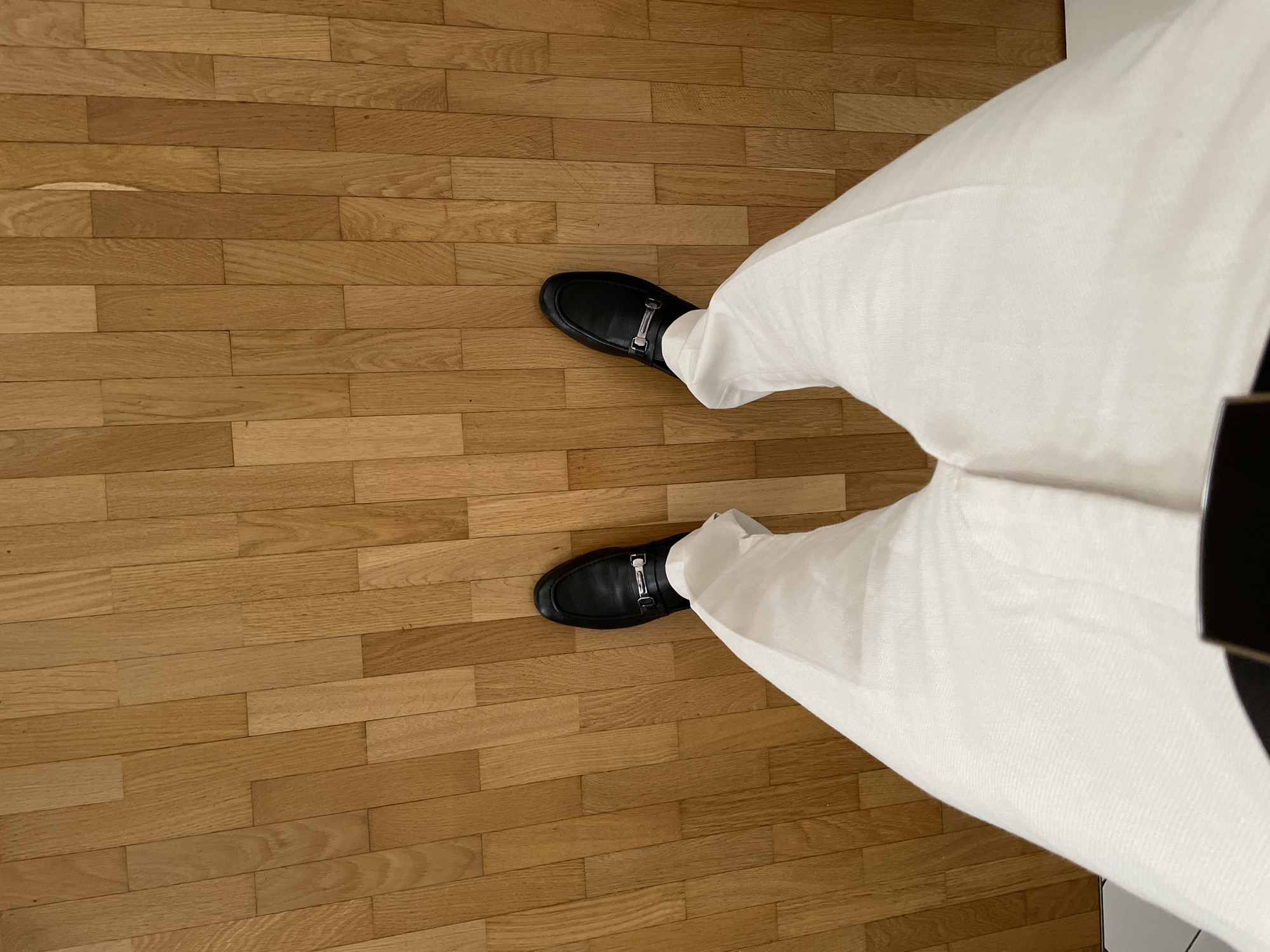 The image shows a person standing on a hardwood floor. The person is wearing white pants and black loafers with silver buckles. The background is a wooden floor and a wall.