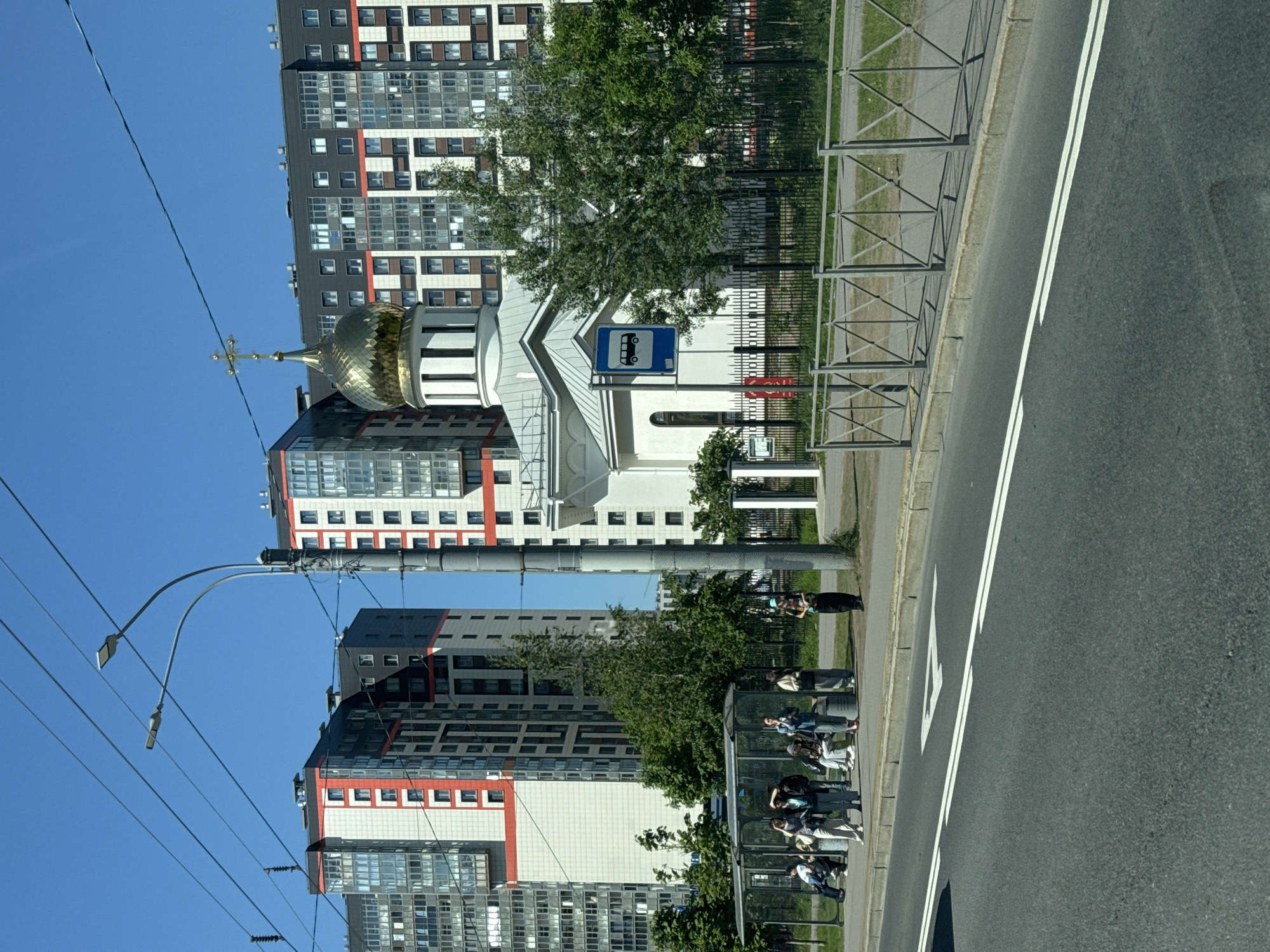 The image, rotated 90 degrees clockwise, presents a daytime street scene in Sinyavino, Russia, under a clear blue sky. In the immediate foreground, a multi-lane asphalt road with distinct white double solid and dashed lines runs alongside a curb. A grey metal barrier fence separates the road from a paved sidewalk and a strip of grass.
On the sidewalk, a modern bus stop shelter is visible, occupied by several people waiting. Some individuals are seated within the shelter, while others stand nearby. Further down the sidewalk, another person can be seen standing.
The background is dominated by a cluster of tall, contemporary apartment buildings. These structures feature multiple stories with grey and white facades, accented by striking red horizontal bands and window frames. Nestled among these modern residences is a white church, distinguished by its gleaming golden, onion-shaped dome topped with an Orthodox cross. A blue sign depicting a white bus icon, indicating a bus stop, is visible near the church. Green trees are interspersed throughout the scene, providing foliage.
Overhead, a streetlight pole and various utility wires crisscross against the bright, cloudless sky. The presence of clear shadows suggests bright sunlight and good weather conditions. The scene captures a blend of modern urban development with traditional religious architecture. No legible text is visible other than the bus icon on the sign.