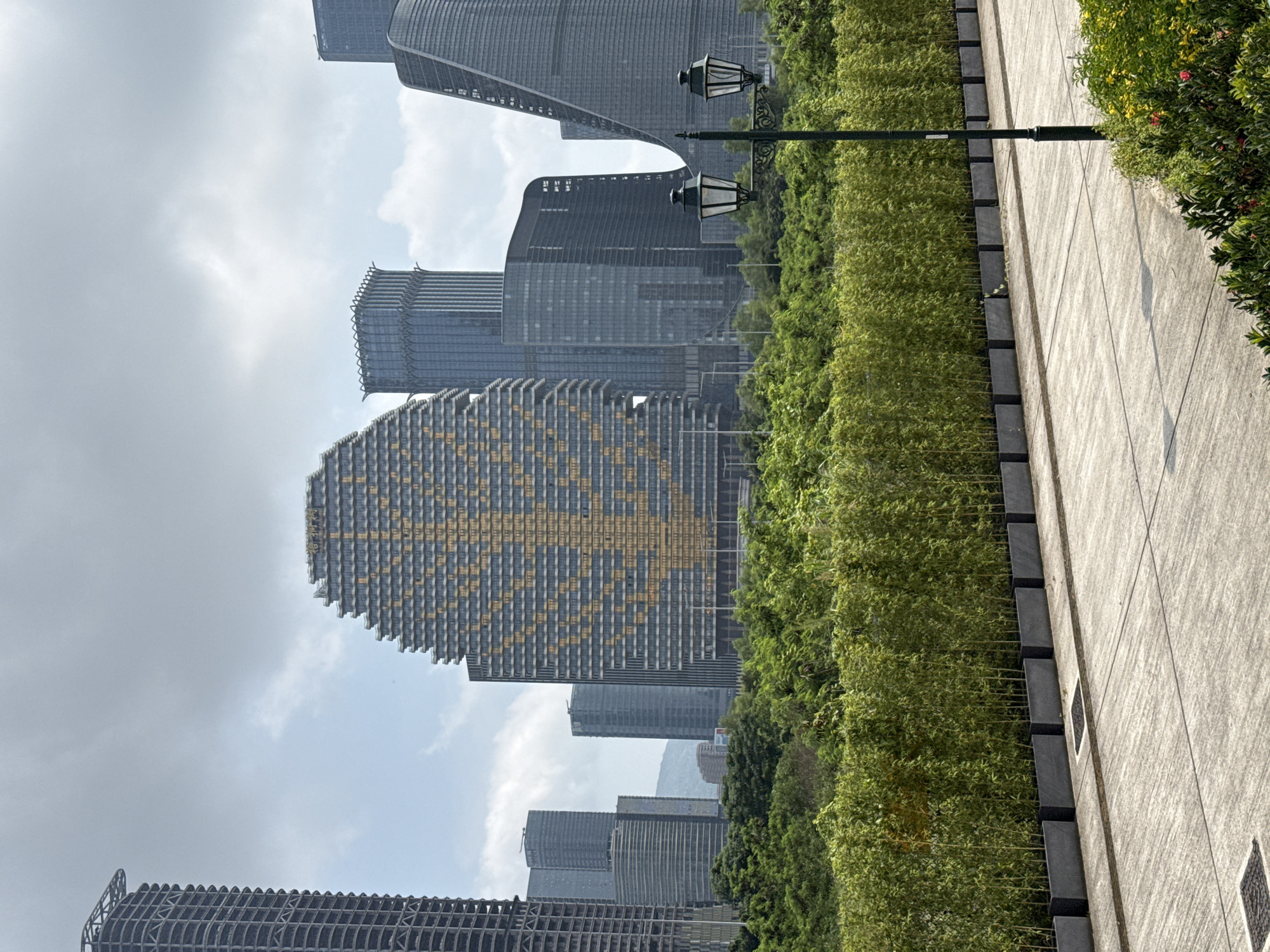 The image depicts a view of the city skyline in Unknown, Macao, featuring modern high-rise buildings with unique architectural designs. The foreground consists of a paved walkway bordered by lush green foliage, including hedges and bamboo. A vintage-style lamppost stands along the walkway. The buildings showcase a mix of materials and facades, including grid-like patterns and curved structures. The sky is overcast with scattered clouds.