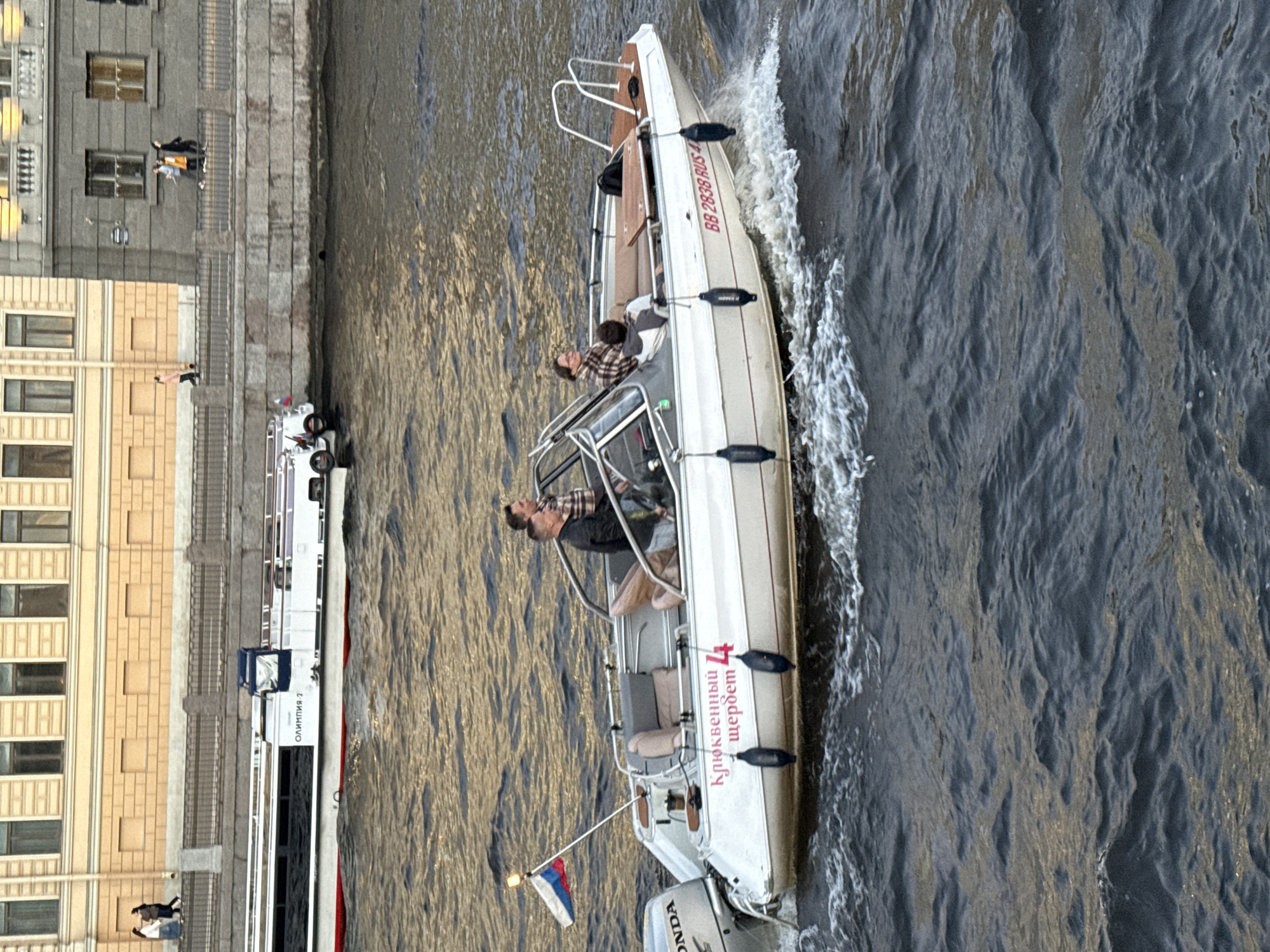 In Saint Petersburg, Russia, a white motorboat with a blue canopy is traveling on the Neva River. The boat is carrying four individuals: two men in the front and two people, a man and a woman, seated further back. The boat has a Russian flag flying from its stern and the registration number "BB 2838 RUS 4" is visible on its side, along with the text "Клюквенный щербет" which translates to "Cranberry Sherbet". The boat is creating a wake as it moves through the choppy, dark water. In the background, a large, light-colored building with numerous windows lines the embankment, and a larger passenger ship is docked or moving slowly in the distance. Several people are visible walking along the embankment. The sky is overcast, and the lighting suggests it is late afternoon or early evening.