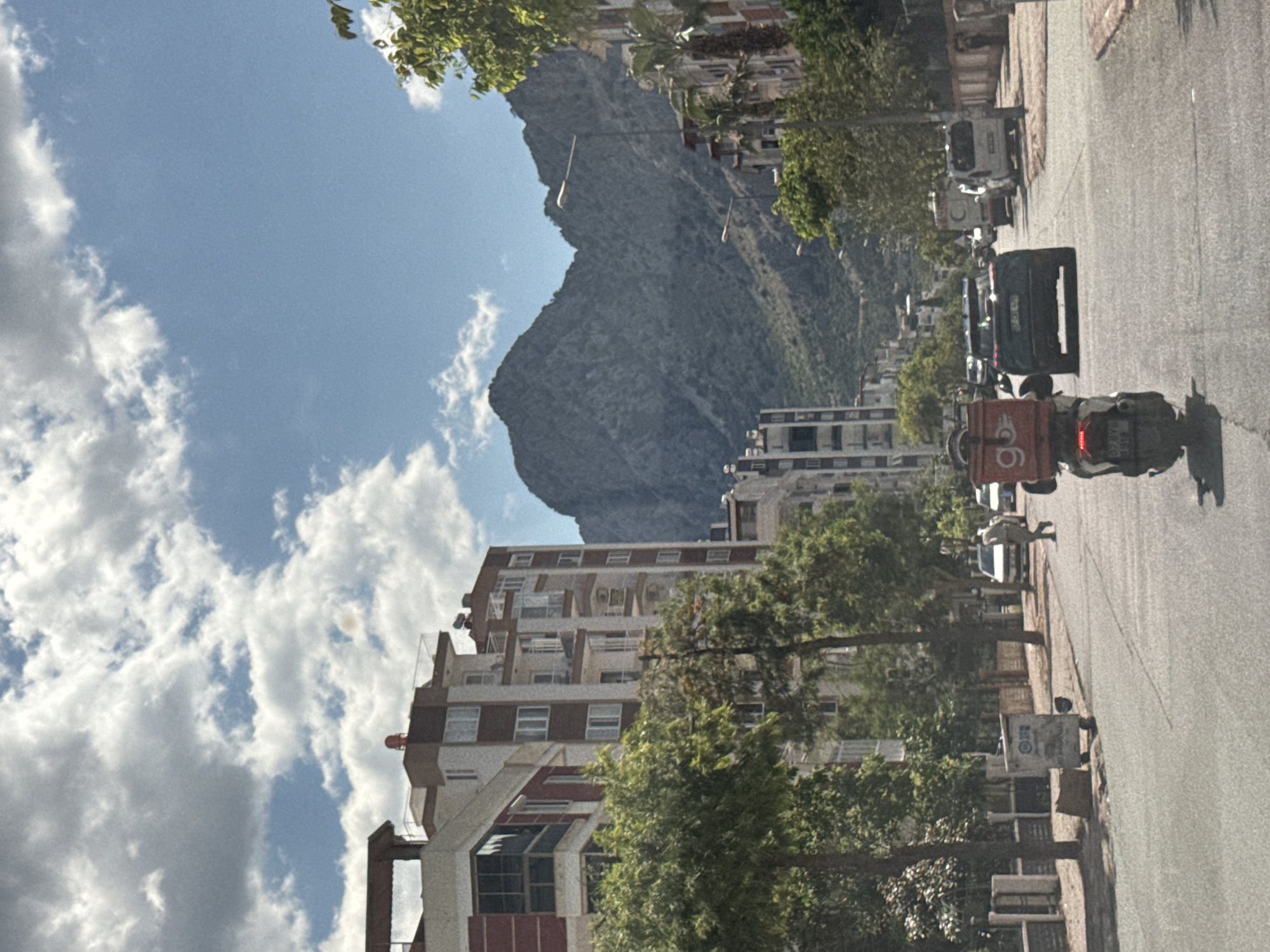 An eye-level photograph captures a street scene in Konyaaltı, Turkey. The foreground features a motorcycle with a delivery box, positioned on a sunlit road. Behind the motorcycle, parked cars, pedestrians, street signs, and trees line the side of the street. The buildings lining the street are of varying heights. In the backdrop is a large mountain range against a blue sky with white clouds.