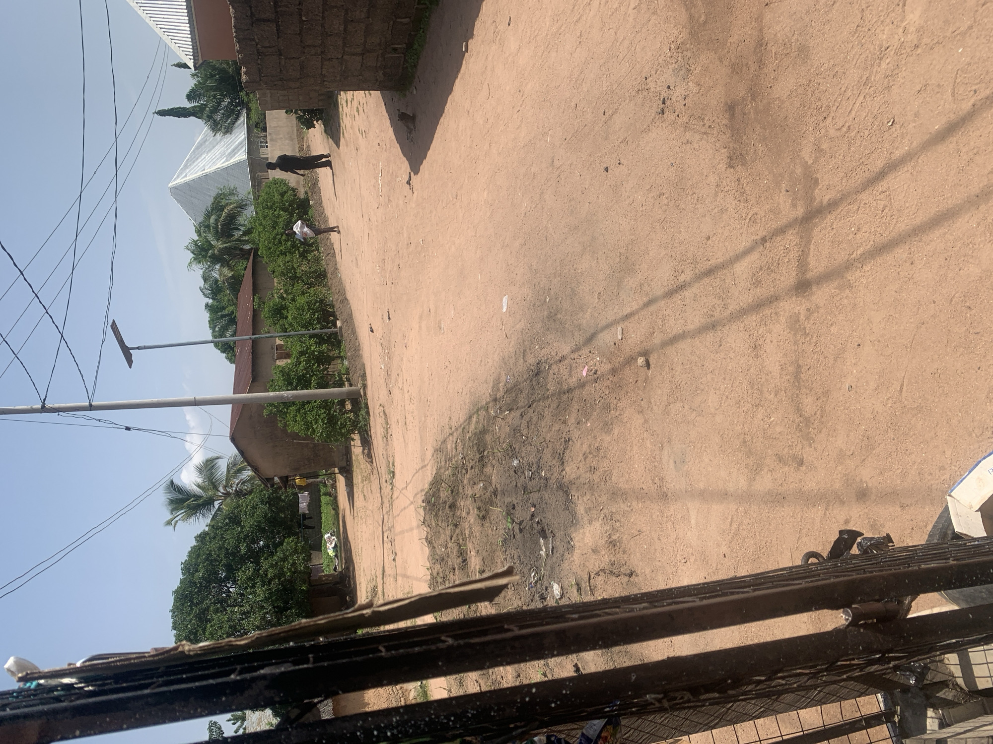 The image depicts a dusty, unpaved road in a rural or semi-urban area, likely Jalingo, Nigeria, under a bright, clear sky. The road is a reddish-brown dirt, with some scattered debris and patches of dried mud. On the left side of the road, a dark metal fence or gate is visible in the foreground, partially obscuring the view. Beyond the fence, there are buildings constructed with what appears to be mud or brick, some with corrugated metal roofs. Trees, including a palm tree, and dense green bushes line the road and surround the buildings. In the middle ground, two individuals are walking on the road. One, a person wearing dark clothing, is walking away from the camera. Another, a person wearing a white shirt and shorts, is facing the camera. Further down the road, a distinctive pyramid-shaped building with a silver, reflective roof stands out. A utility pole with electrical wires stretches across the upper part of the image. The overall scene suggests a warm, sunny day in a developing area.