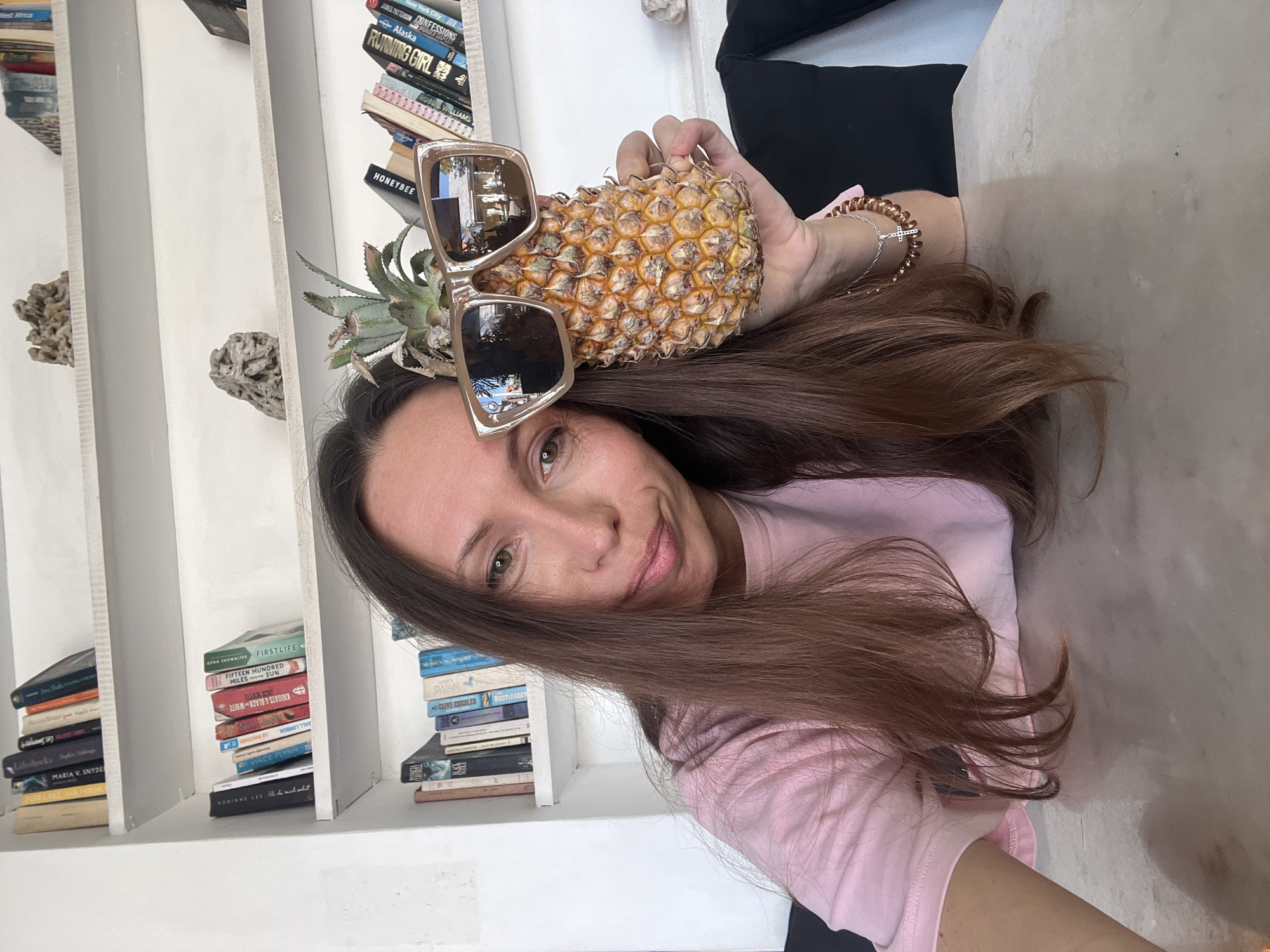 A woman is holding a pineapple on her head with sunglasses on it. She is wearing a pink shirt and has long brown hair. Behind her is a white bookshelf filled with books. Some of the books are "New York City", "Firstlife", "Running Girl", "Confessions", "Bootlegger", "Swan" and "Honeybee". There are also coral decorations on the shelves. The location is likely indoors, possibly in North Kuta, Indonesia based on the writing on a book.