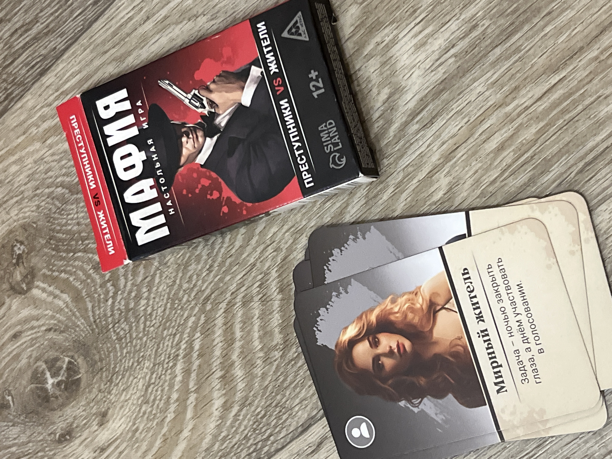 The image depicts a close-up, slightly overhead shot of a table with two items from the game "Mafia" or "Мафия". On the right is the game box, featuring a man in a suit and hat holding a pistol against a red, splatter-like background. The box has text in Russian, including "МАФИЯ", "НАСТОЛЬНАЯ ИГРА", and "ПРЕСТУПНИКИ VS ЖИТЕЛИ". To the left of the box are several cards from the game, fanned out and overlapping. The visible card prominently displays an illustration of a woman with blonde hair and a white circular icon with a silhouette of a person. Below the illustration, text in Russian reads "Мирный житель" (Peaceful resident) and describes the character's task. The items are placed on a wooden surface with a visible wood grain pattern. There are no identifiable landmarks or specific details that would suggest a location in Дачное, Russia.