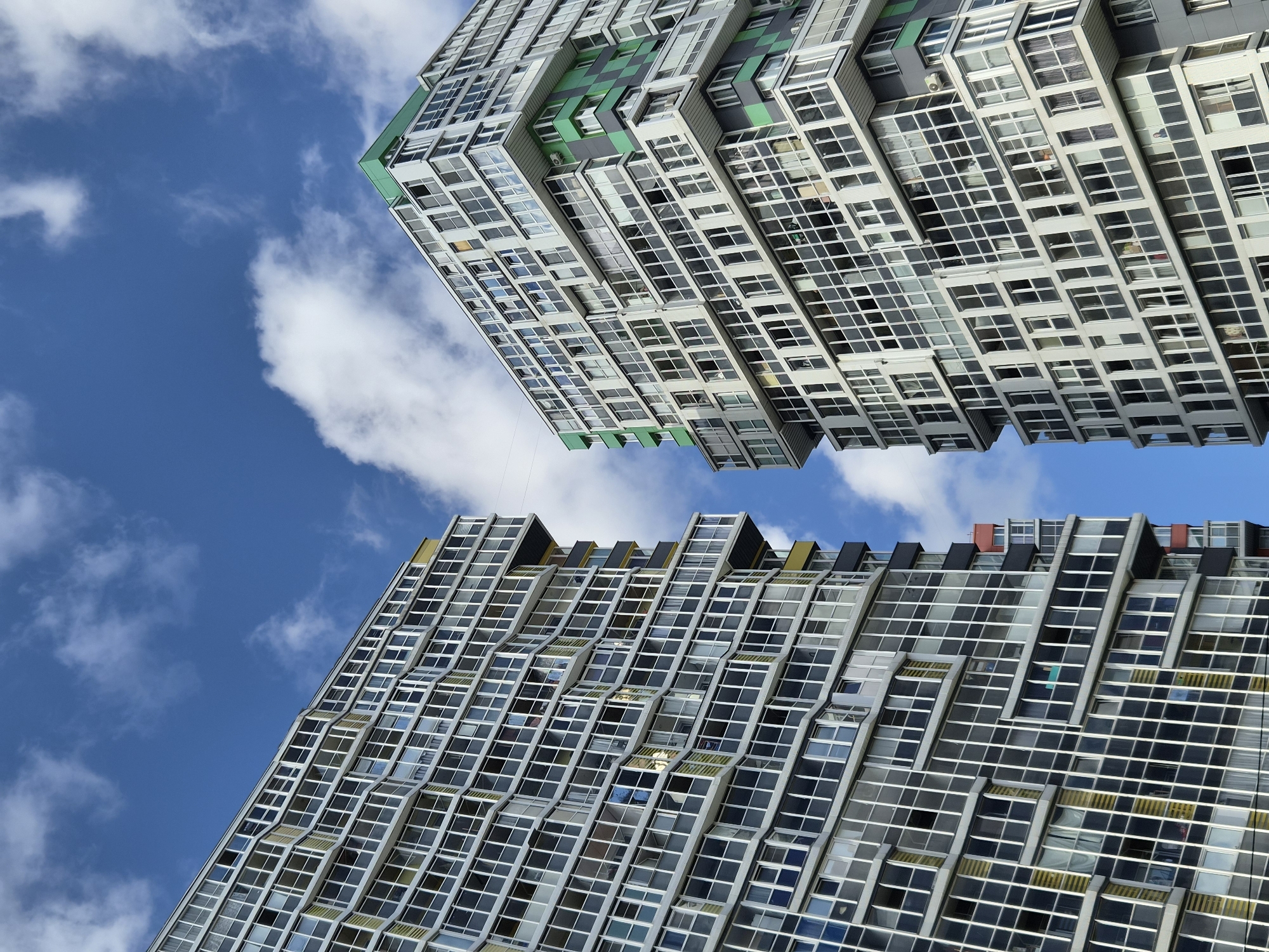 In Kudrovo, Russia, two modern apartment buildings rise against a bright blue sky dotted with white clouds. The building on the left is characterized by a predominantly gray facade with numerous windows and balconies. Yellow accents are visible on some of its balconies. The building on the right features a similar structure with a multitude of windows and balconies, but it incorporates vibrant green panels and a green-tiled roof, adding a distinct color contrast. The perspective is from below, looking up at the towering structures, emphasizing their height and architectural design. There are no visible people or ongoing activities in the scene.