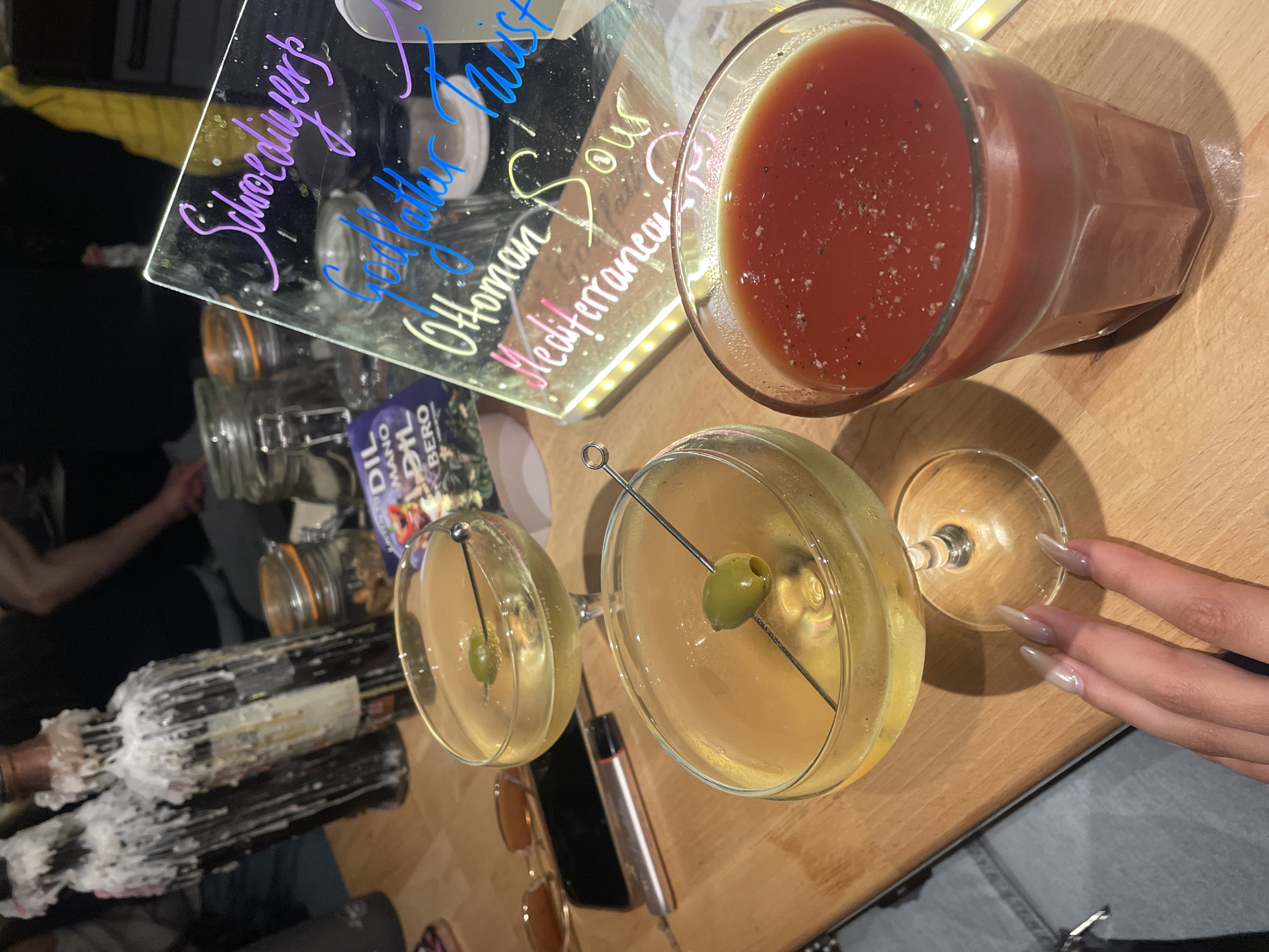 An overhead shot captures a cozy, dimly lit social setting in Sofia, Bulgaria, likely during the evening. The main focus is on a collection of drinks and personal items resting on a light-colored wooden surface, possibly a table or bar counter.
In the foreground, two martini glasses each hold a clear, yellowish cocktail garnished with a single green olive on a skewer. To their right, a hand with long, manicured nails is visible. Between the martini glasses, a black smartphone and a rose-gold vape pen are placed. A pair of sunglasses with brown lenses is also nearby.
Above the martini glasses, a tall glass contains a reddish beverage, possibly a Bloody Mary, topped with visible pepper flakes. Behind these drinks, a clear, rectangular acrylic sign with handwritten text in various neon colors is illuminated from beneath, suggesting it's a menu or specials board. Visible text on the sign includes "Schoedlinger" (in purple), "Godfather Twist" (in blue), "Ottoman Sour" (in yellow), and "Mediterranean" (in pink).
To the left of the illuminated sign, two dark-colored bottles, heavily encrusted with melted white wax, are positioned, implying their use as candle holders. Behind these bottles, two glass jars with orange clamp-tops are partially visible. A small package with the words "OIL MANO" and "BERO" printed on it can be seen behind the jars. In the background on the far left, a person's arm and shoulder are indistinctly visible. The overall scene suggests a relaxed, social gathering where drinks are being enjoyed.
