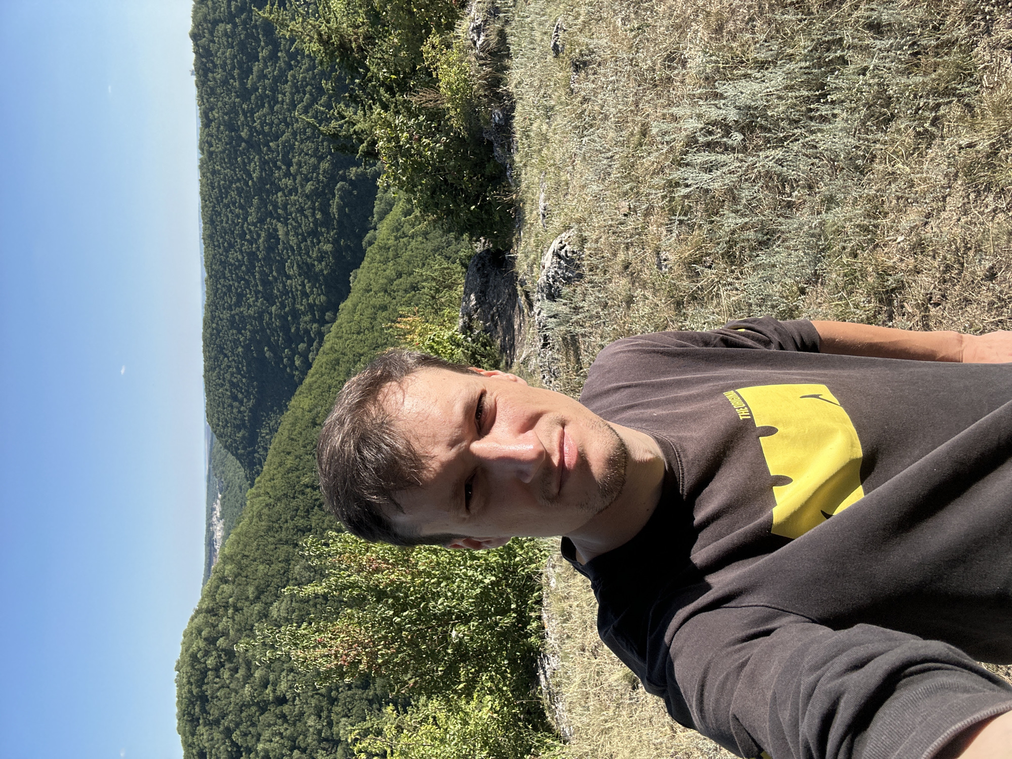 A selfie is taken by a man with short brown hair and stubble, wearing a dark long-sleeved shirt with a large yellow smiley face design on the left chest and the text "THE ORIGINAL" above it. He is standing on a grassy, rocky hill overlooking a valley filled with lush green trees. The sky is clear blue. In the distance, faint buildings can be seen nestled amongst the trees, possibly indicating the location of Pryvorottia Druhe, Ukraine. The sun is casting shadows, indicating it is daytime.