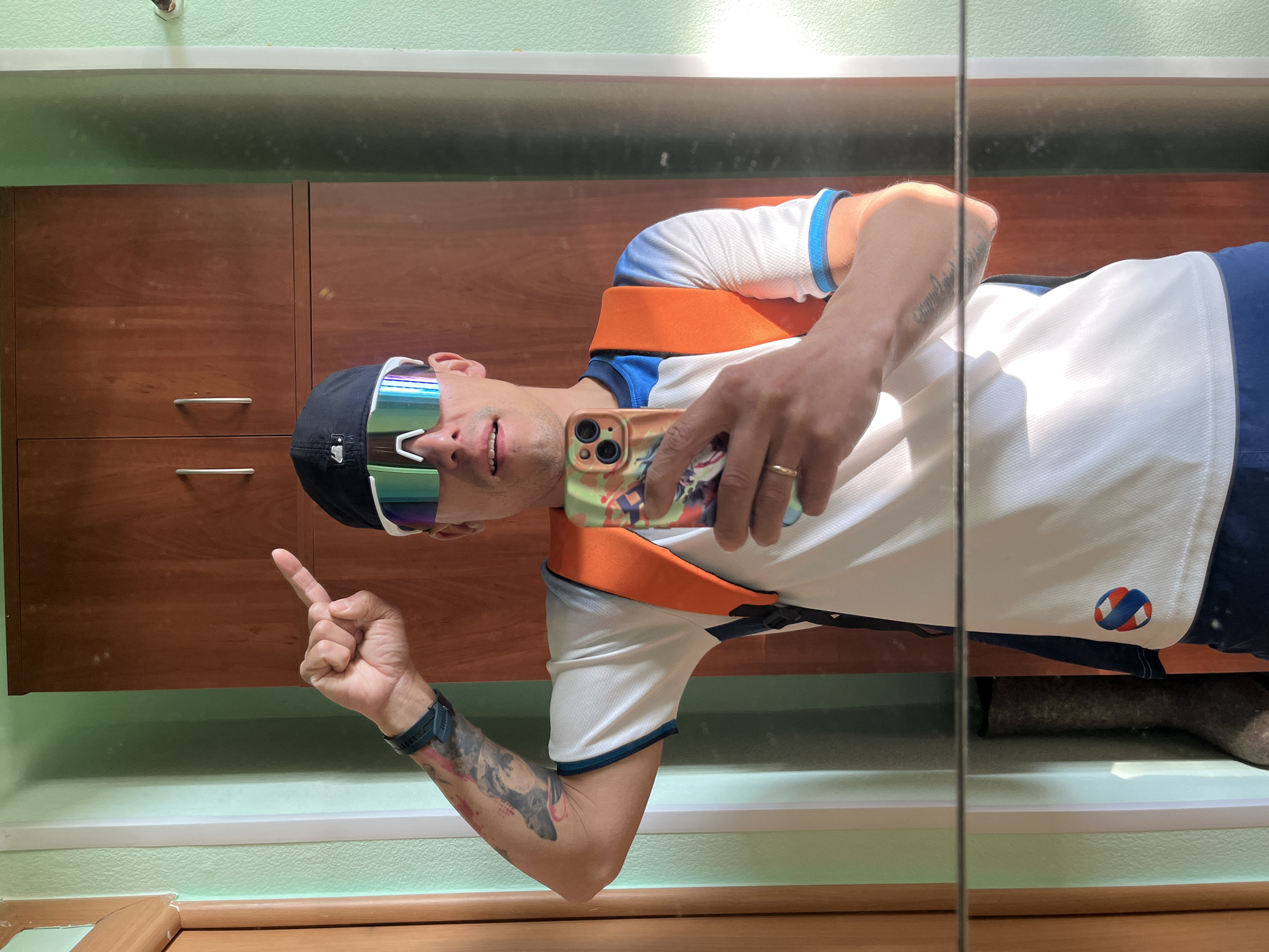 A man in a white and blue-trimmed t-shirt, black baseball cap, and reflective sunglasses is taking a selfie in a mirror, pointing upwards with his left index finger. He is holding a smartphone in his right hand, which has a colorful case featuring a cartoon character. Tattoos are visible on both arms. He wears a watch on his left wrist and a ring on his right ring finger. An orange strap runs across his torso.
The background includes light green walls, a dark wood-paneled cabinet with silver handles, a white trim, and a glimpse of a tan boot at the bottom.
The location is in an interior of a building, but there is no further information in this image to indicate whether it is located in the city of Chaikovsky, Russia.