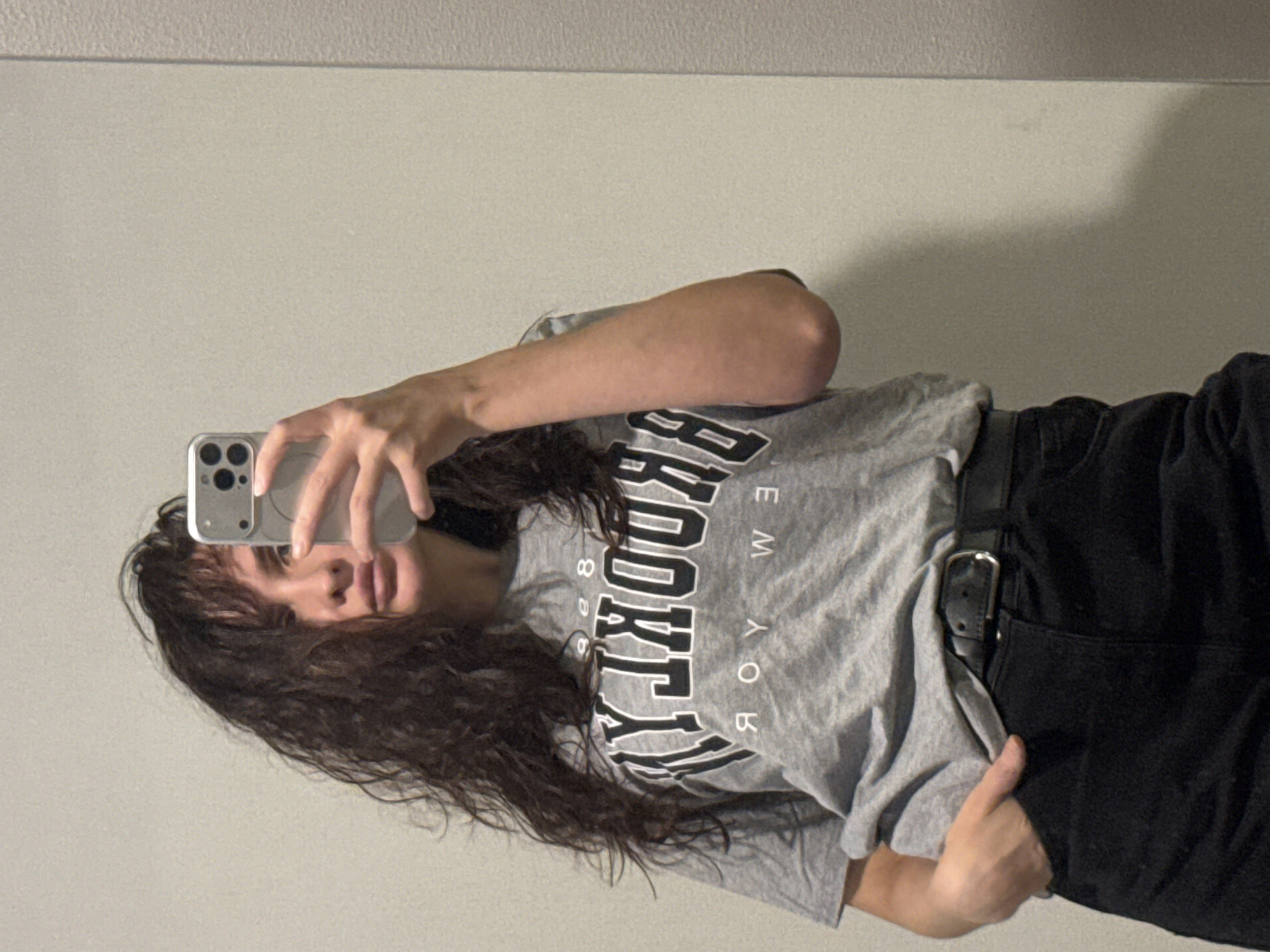 A young woman takes a selfie in front of a blank wall, likely indoors. She is wearing a gray t-shirt with the word "Brooklyn" printed in large, black letters, black pants with a belt, and has long, curly brown hair. She is holding a silver iPhone in her right hand, using it to take the photo. The exact location is not discernible from the image; the city of Chaikovsky, Russia, cannot be confirmed.