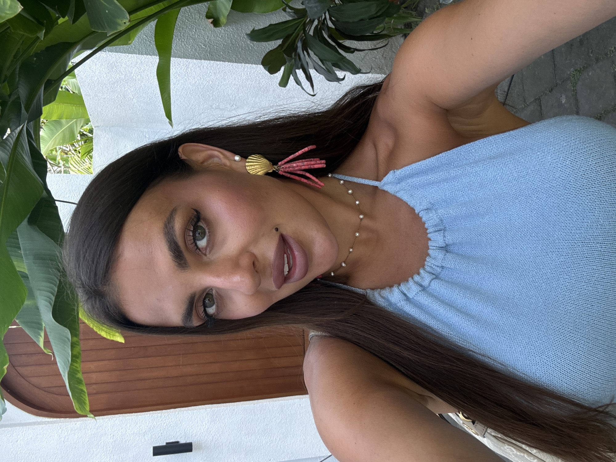A woman with long, dark hair is posing for a selfie. She is wearing a light blue top and decorative earrings, and a delicate pearl necklace. The background includes foliage and a white wall, suggesting an outdoor location. The location may be North Kuta, Indonesia.