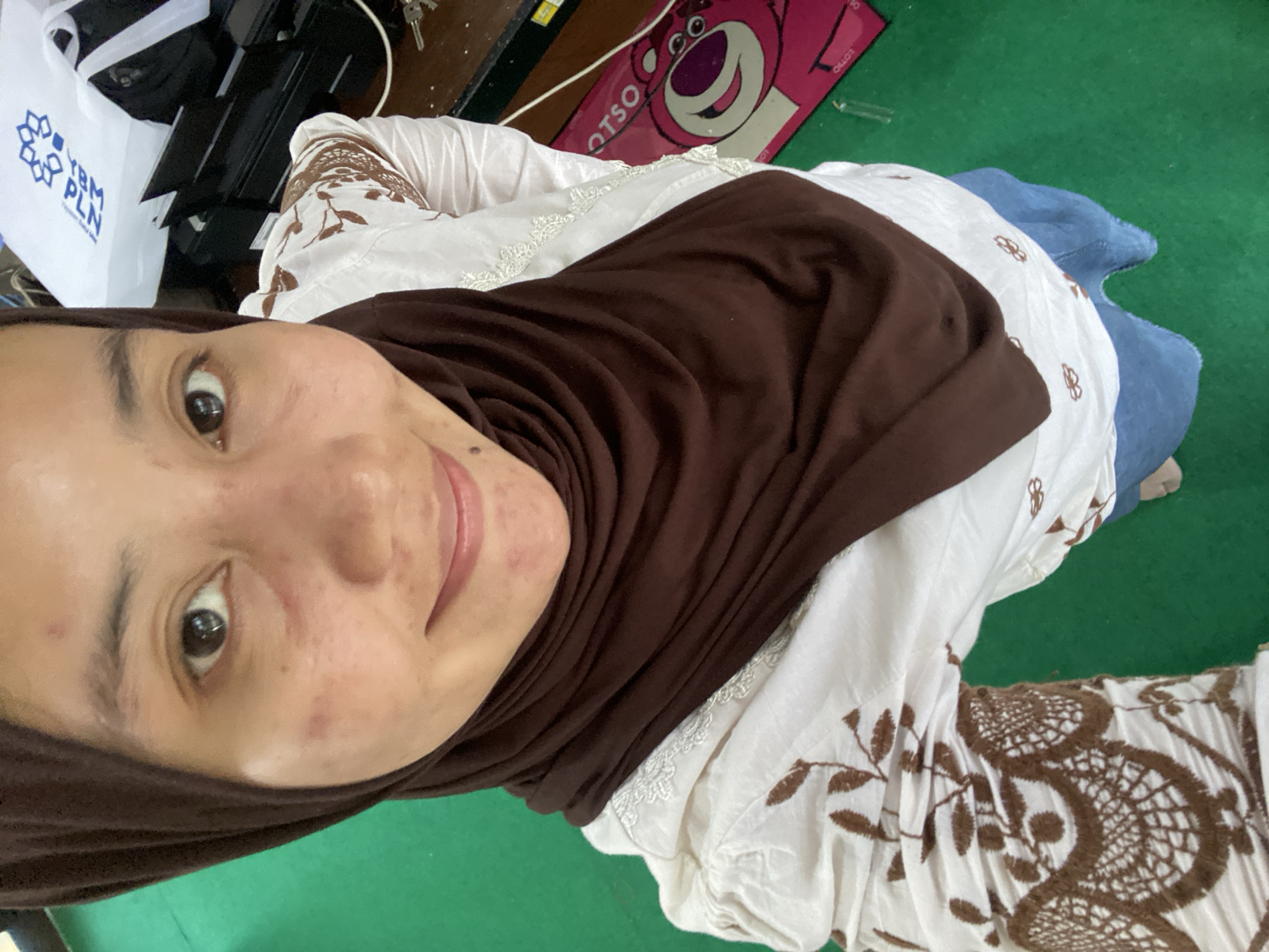 A woman wearing a brown hijab and a white shirt with brown embroidery is taking a selfie in what appears to be an office setting. She has a light complexion and some blemishes on her face. The background includes office equipment such as a printer, a white bag with a blue logo, and a pink mat with a cartoon character on it. The floor is green. The image was taken in Banjarbaru, Indonesia.