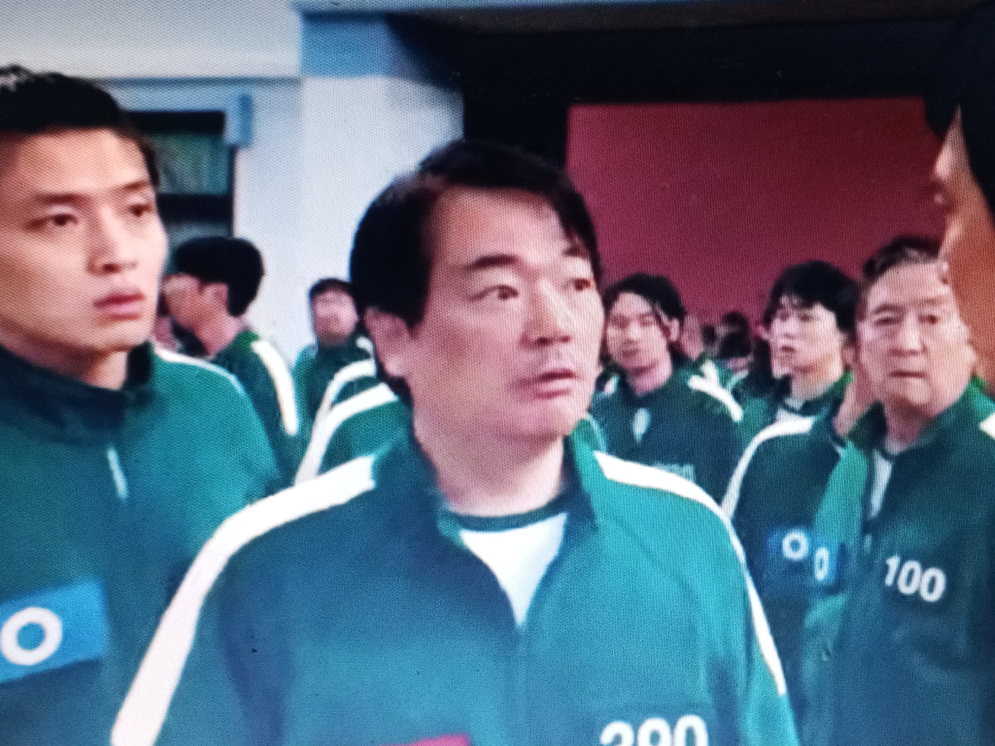 The image depicts a group of people wearing green tracksuits with white stripes, arranged in a crowded indoor space. Numbers are visible on the tracksuits. The people are of East Asian descent and appear to be looking in different directions. A wall is visible in the background with a red section. The location is Unknown, Unknown.