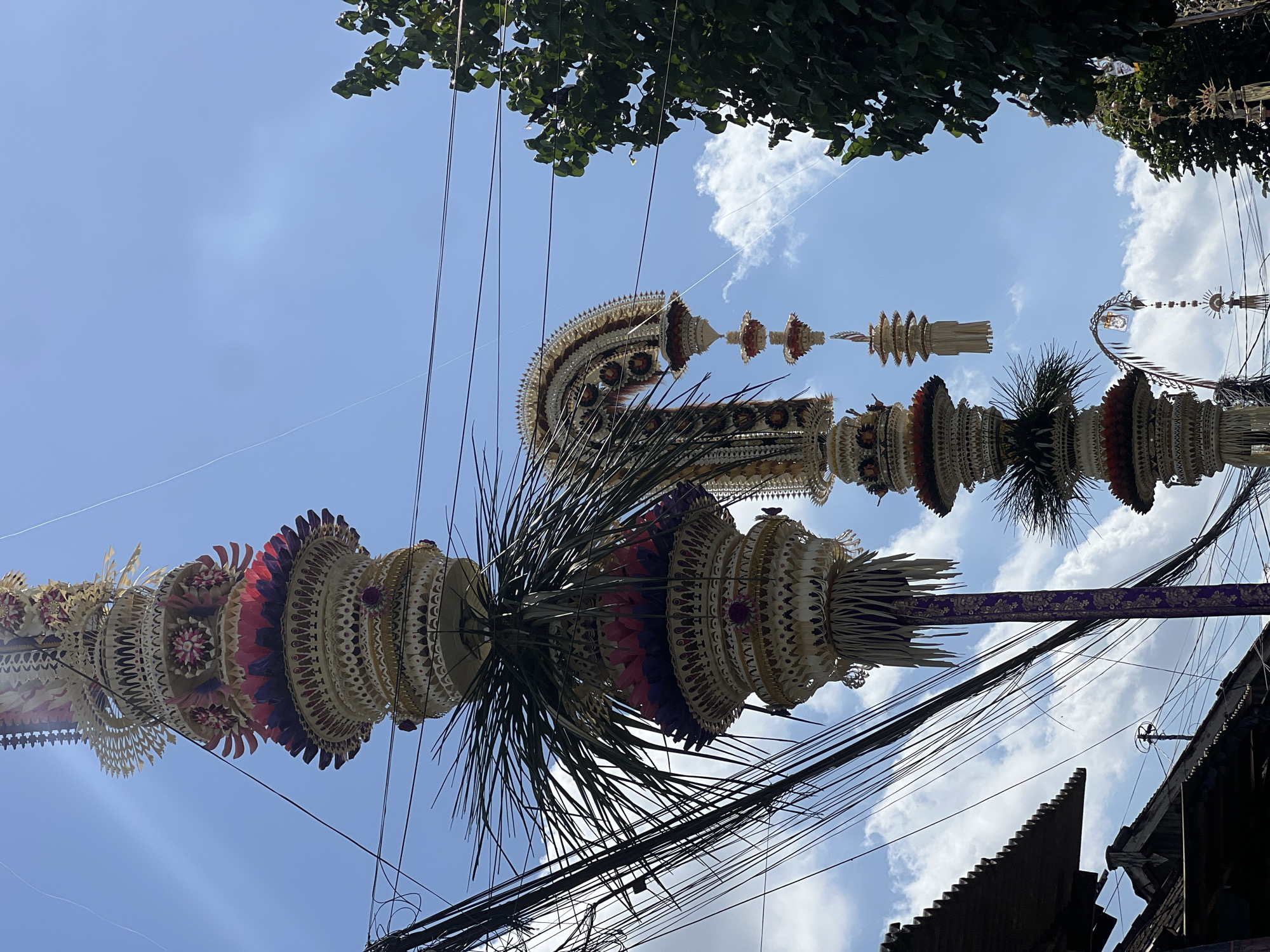 The image depicts a street scene in North Kuta, Indonesia, dominated by elaborate, towering Balinese offerings known as "Penjor." These decorative poles, adorned with intricate woven palm leaves, flowers, and fruits, are erected for religious ceremonies, often associated with the Galungan and Kuningan festivals. The Penjor are positioned against a bright blue sky dotted with white clouds, with some partially obscured by dense overhead power lines and a lush green tree on the right side of the frame. Traditional Balinese architecture, characterized by dark, slanted roofs, is visible in the lower left corner of the image. The composition suggests a cultural event is taking place or has recently occurred, as indicated by the presence and scale of the Penjor.