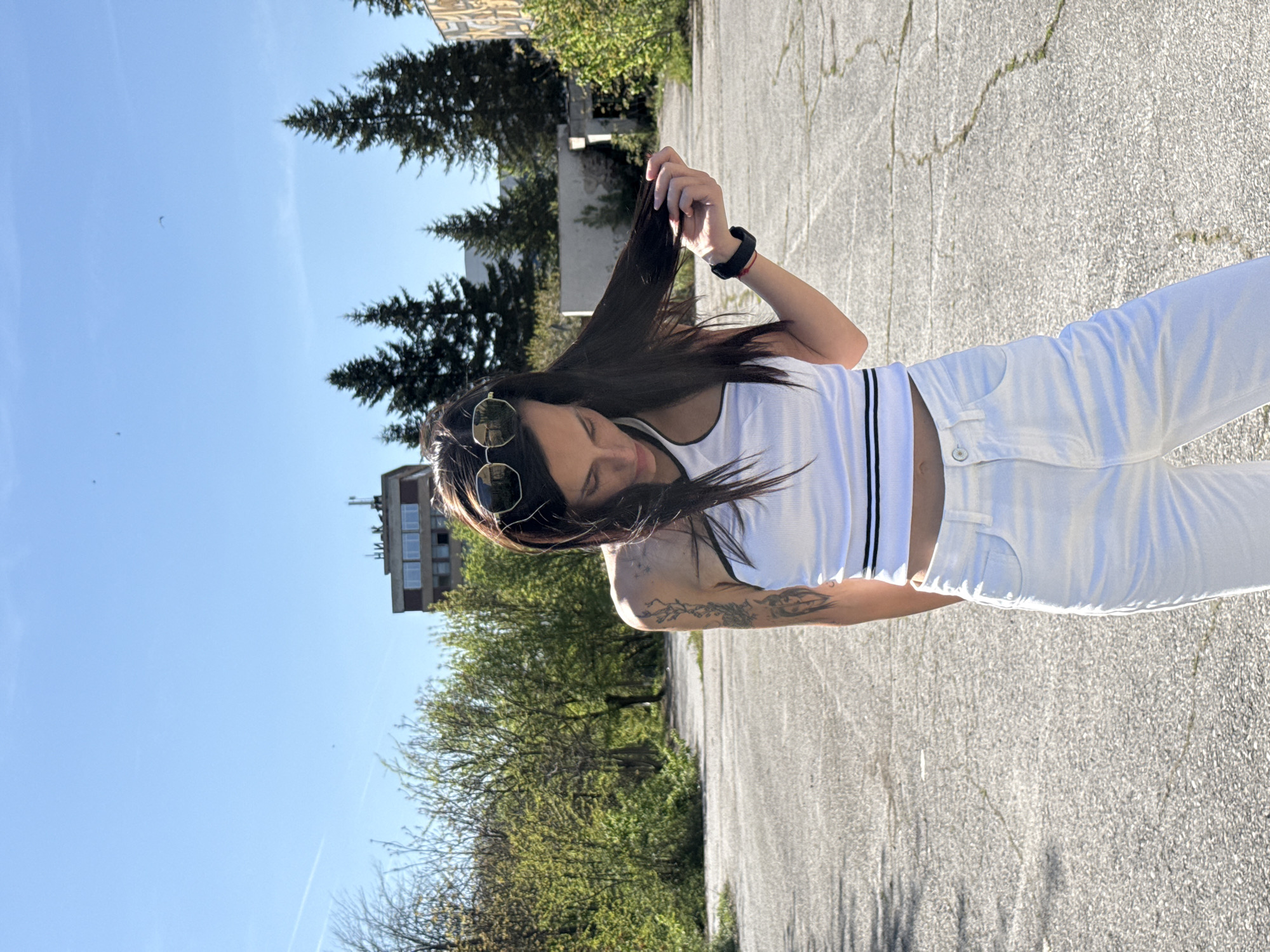A young woman stands on cracked concrete, facing the viewer and looking down. She is wearing white jeans and a white tank top with black stripes. She has long, dark hair, and sunglasses are perched on her forehead. Her left arm has a tattoo. She is holding a section of her hair with her right hand.
Behind her are trees and buildings. The sky is blue and mostly clear. The location is likely in an urban area in Pleven, Bulgaria, based on the architecture and environment. The scene suggests a casual outdoor setting.
