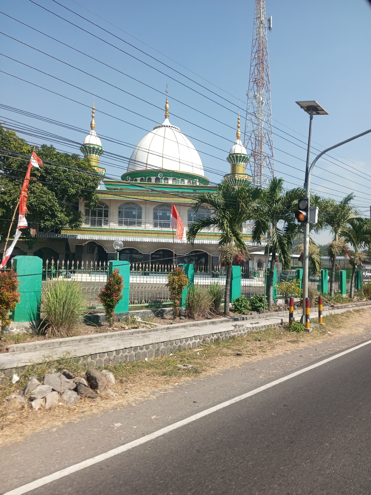 The image shows a mosque in Sengon Agung, Indonesia, on a clear, sunny day. The mosque features a large white dome with green trim, two smaller green and gold minarets, and an elaborate green and white facade with arched windows. In front of the mosque is a green fence, and to the left, two Indonesian flags are displayed on poles. Several palm trees and other lush green vegetation are visible around the mosque. A tall telecommunications tower is prominent in the background. A traffic light is also visible to the right, with the amber light illuminated. The foreground shows a paved road with a white line marking its edge, and to the left, a strip of grass, stones, and dry vegetation. The scene appears to be captured from a moving vehicle on the road.