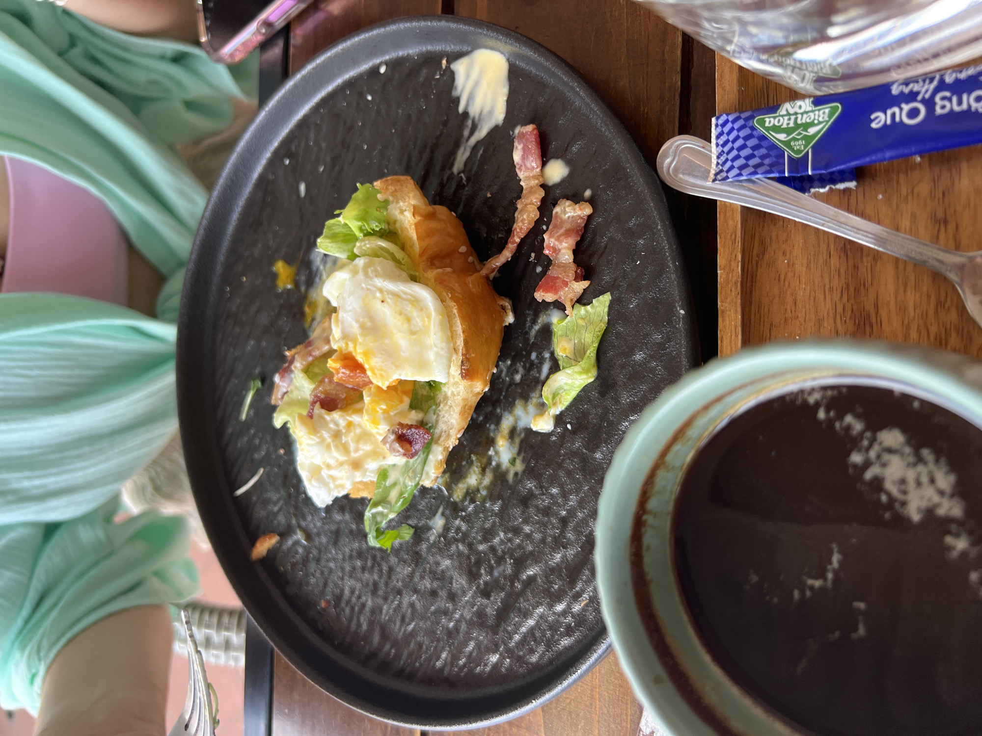 The image captures a close-up, slightly tilted view of a breakfast or brunch setting at what appears to be a cafe or restaurant, indicated by the wooden table and tableware. The primary subject is a partially eaten croissant sandwich served on a dark, textured plate. The sandwich contains a fried egg, bacon, and lettuce, with some sauce or mayonnaise visible on the plate. Next to the plate, a light blue-green cup holds a dark beverage, likely coffee or tea.
Part of a person is visible in the frame, wearing a light green top over a pink garment, with an arm and shoulder area in the upper left. A fork is also partially visible near the bottom left, suggesting the person is mid-meal.
Notable details include a blue and white sugar packet resting on the wooden table next to a spoon. The sugar packet has the brand "Bien Hoa" with "Est. 1969" and Vietnamese text "Đường Quê". A small portion of a phone with a purple case is also visible in the upper right background. The general ambiance suggests a relaxed meal. The location is contextualized as Bac Nha Trang Ward, Vietnam, aligning with the Vietnamese text on the sugar packet.